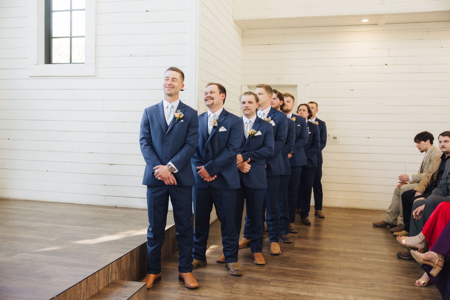 Groomsmen in matching navy suits and brown shoes stand in a line at a wedding ceremony venue.