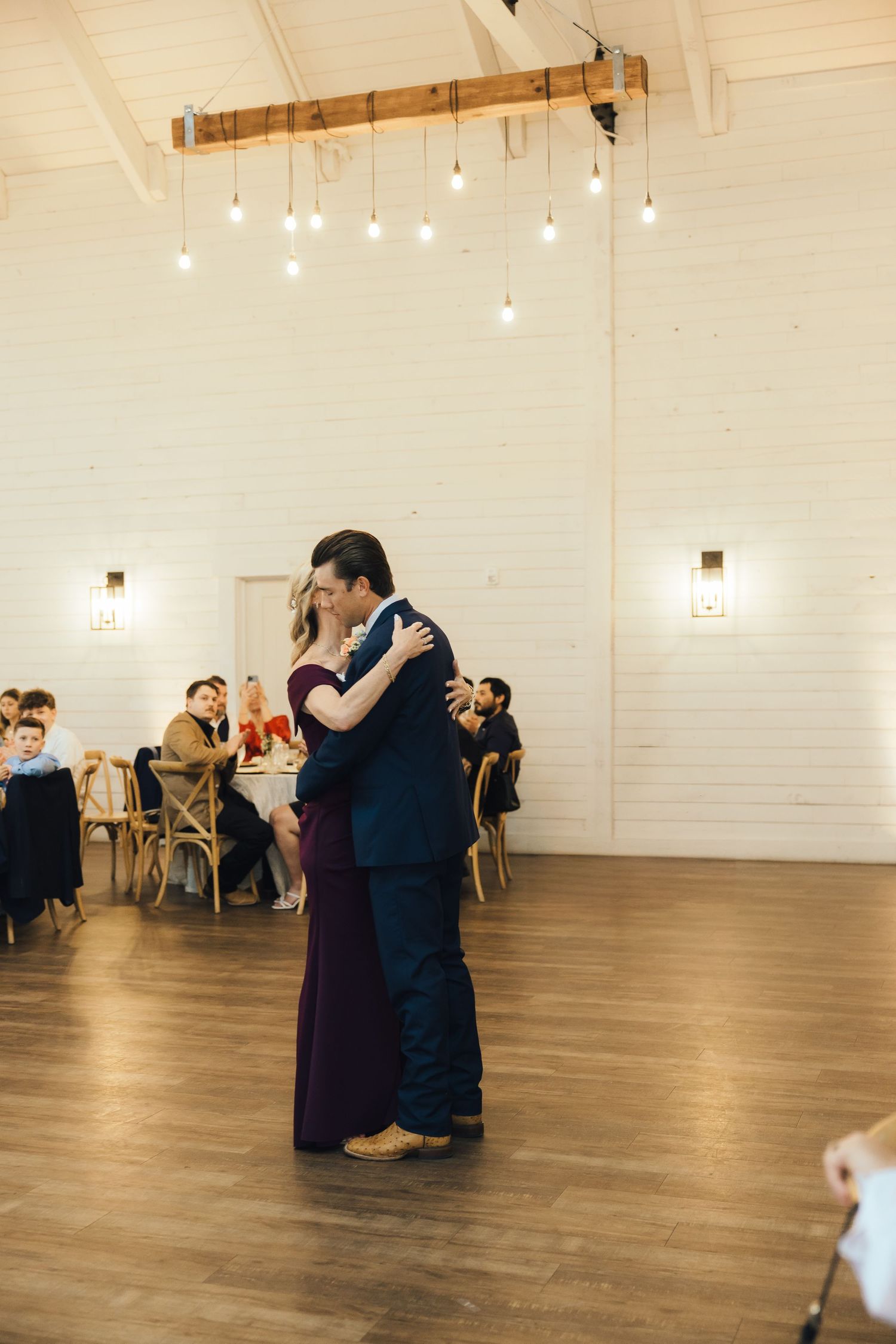 A couple shares a romantic dance moment under hanging lights in an elegant reception venue with wooden floors.