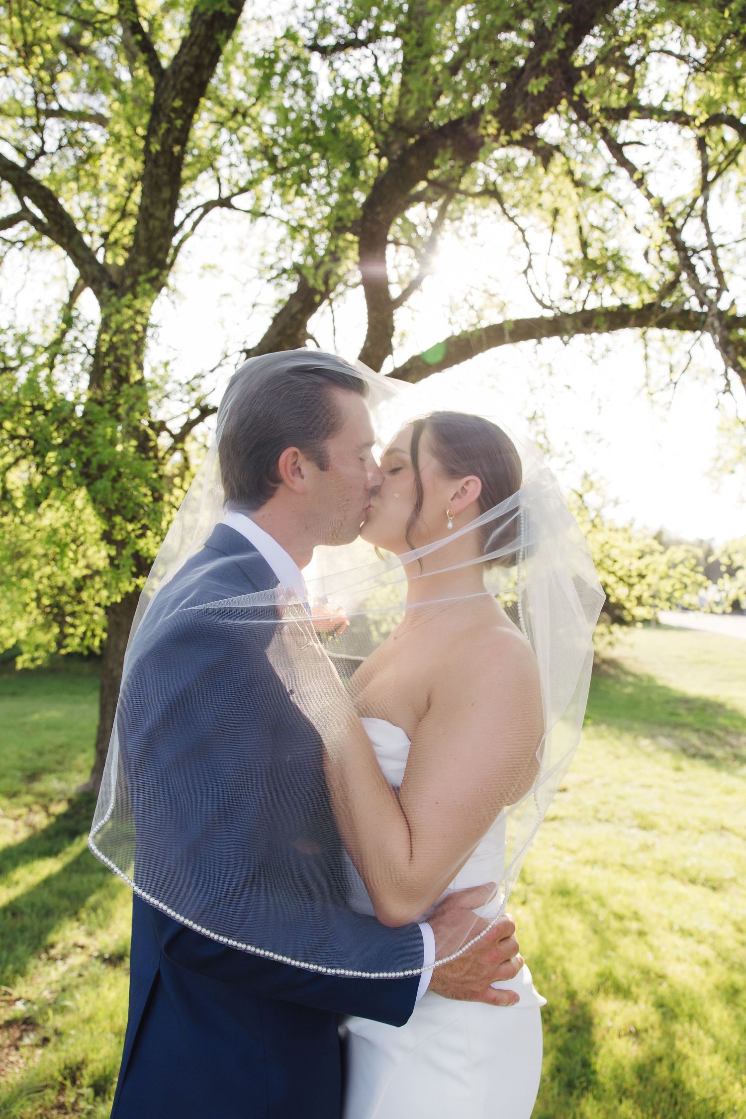 Newlyweds share a romantic kiss outdoors under a sunlit tree during their wedding ceremony.