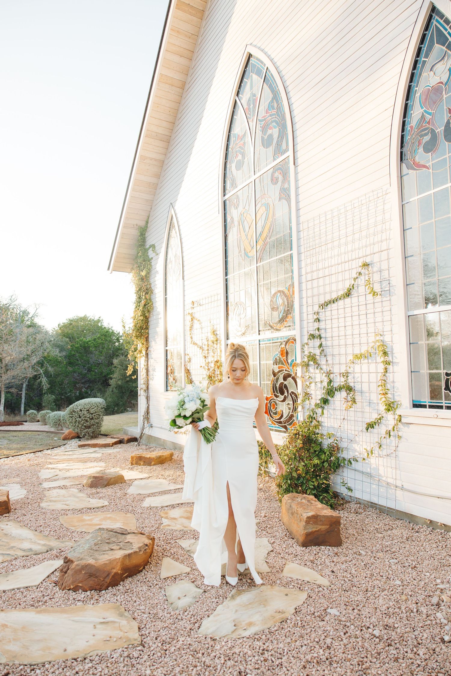 Elegant wedding dress figure stands by white chapel with arched stained glass windows and stone path at golden hour.