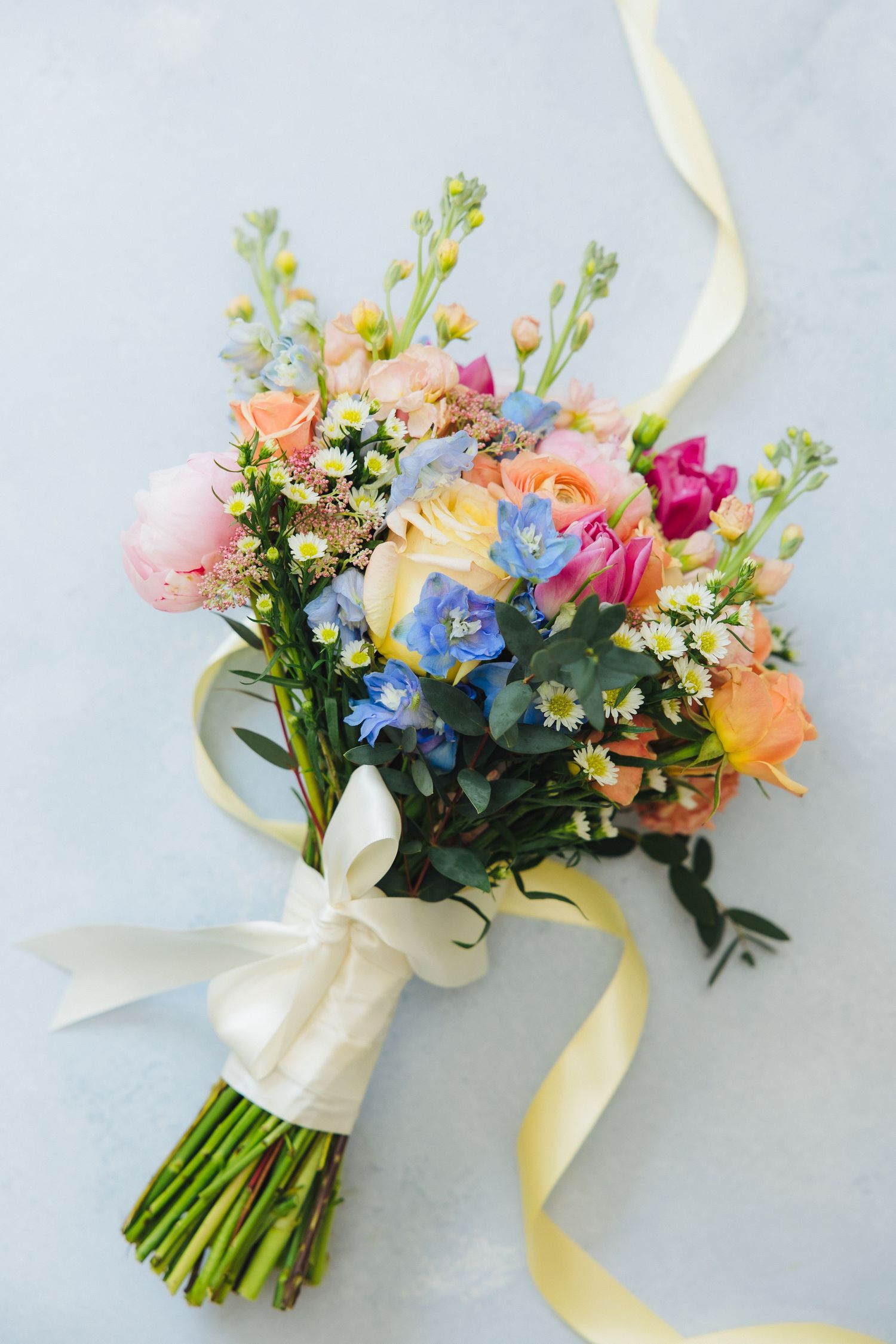 A delicate bridal bouquet featuring pink, peach and blue flowers tied with an ivory ribbon against a light background.