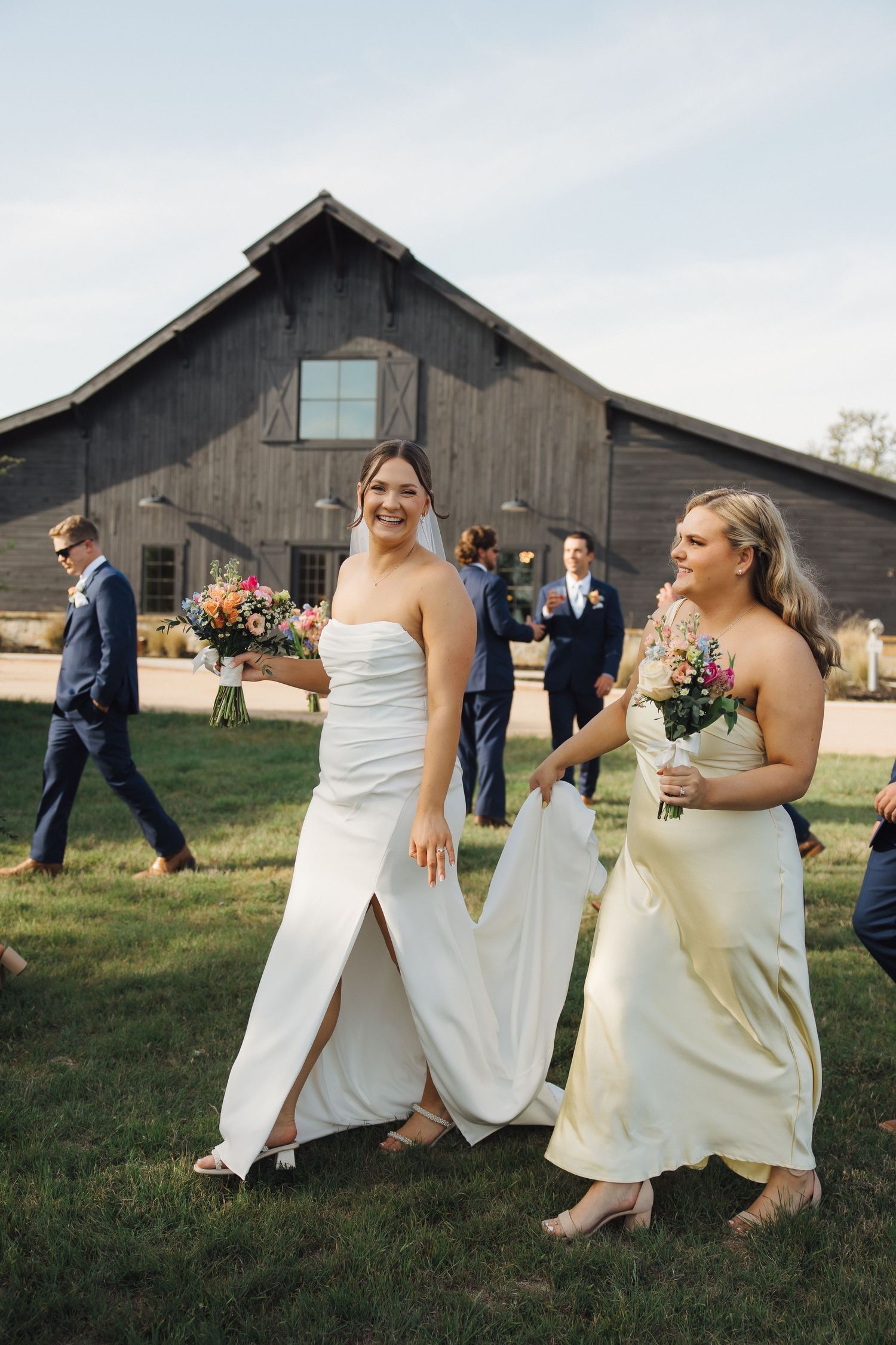 Wedding party members walk on grass outside a rustic barn venue carrying colorful bouquets.