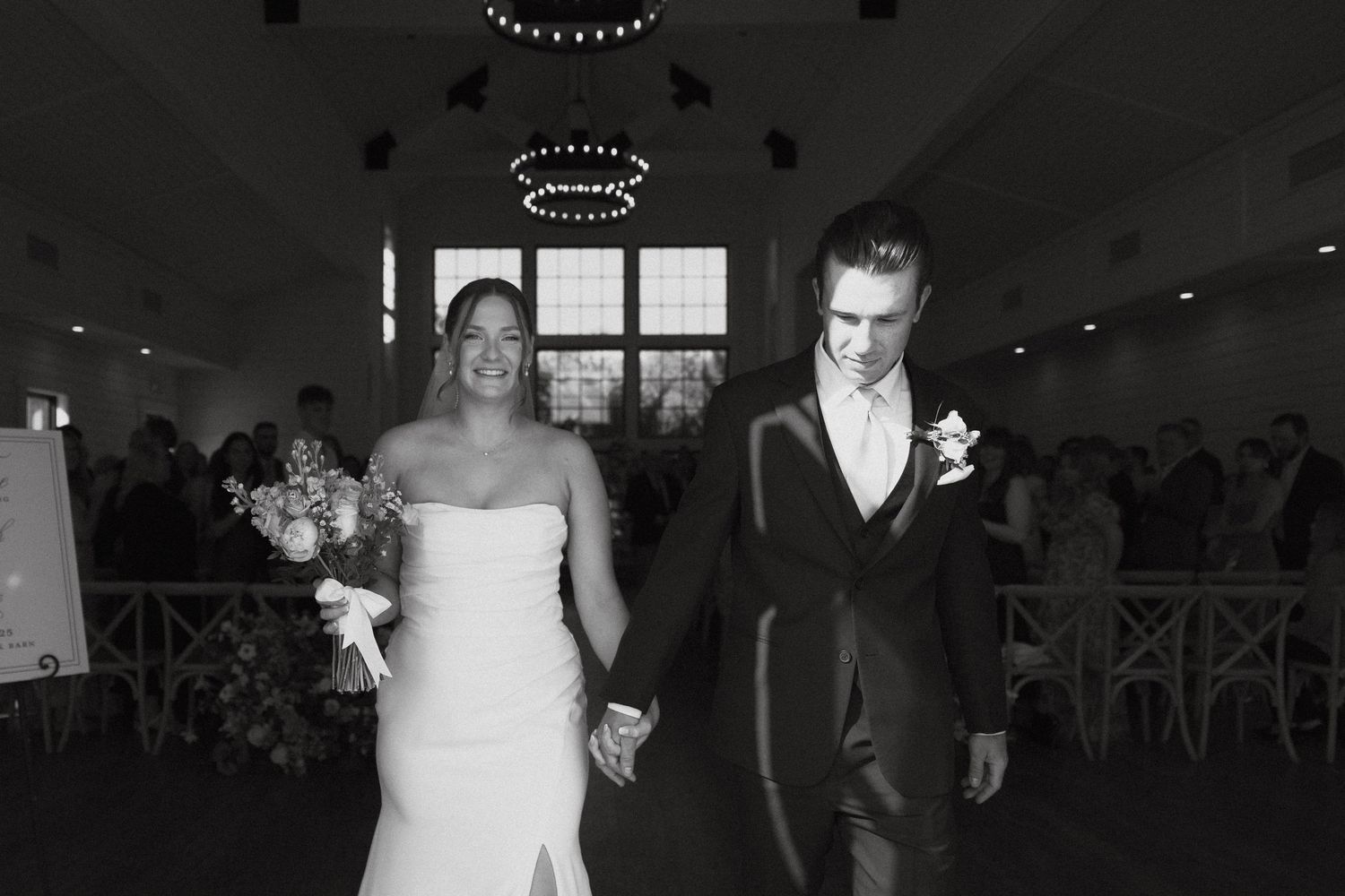 A couple walks down the aisle holding hands in a classic black and white wedding ceremony photo.