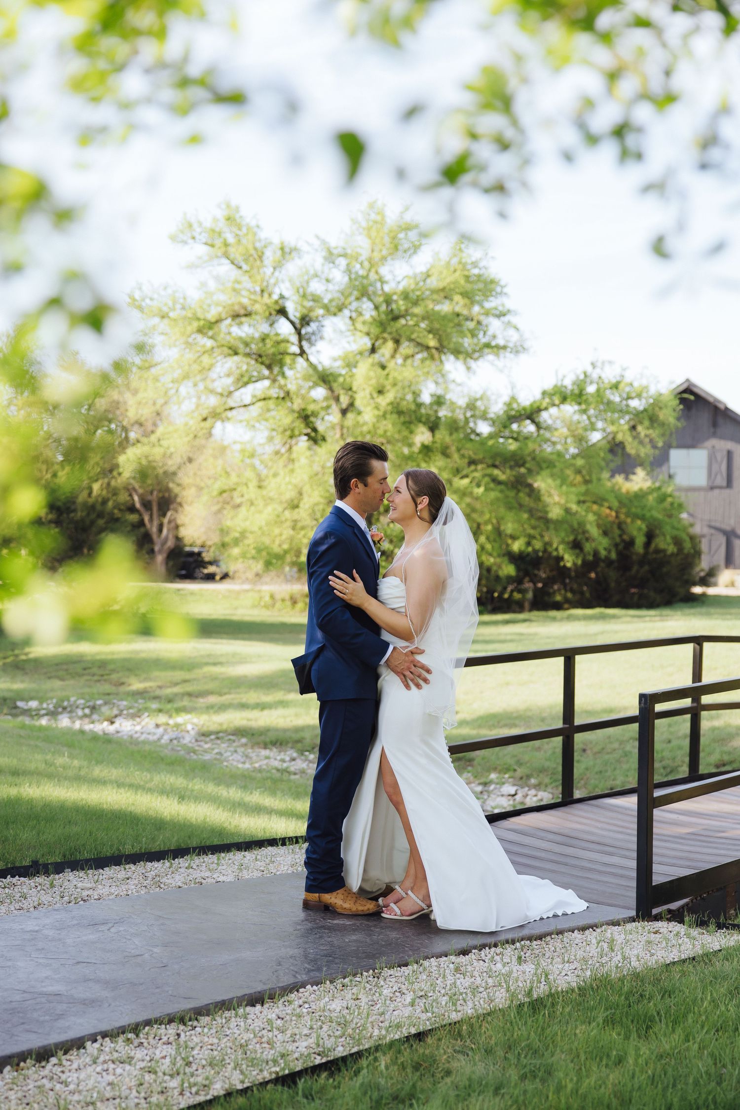 A couple embraces on a wooden bridge walkway surrounded by green trees and landscaping on their wedding day.