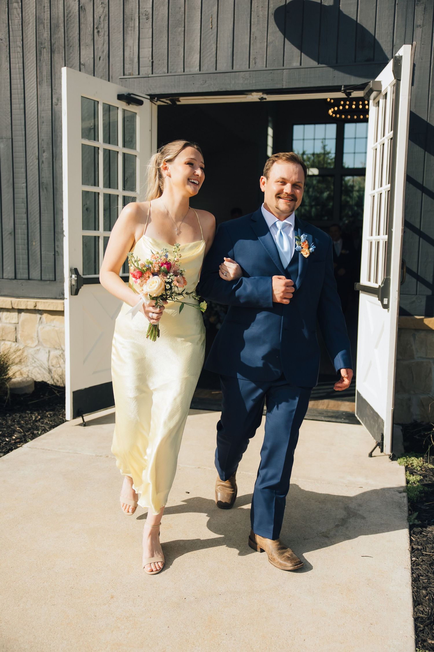 Bride in yellow dress walking with companion in navy suit outside building with white trim and gray siding.