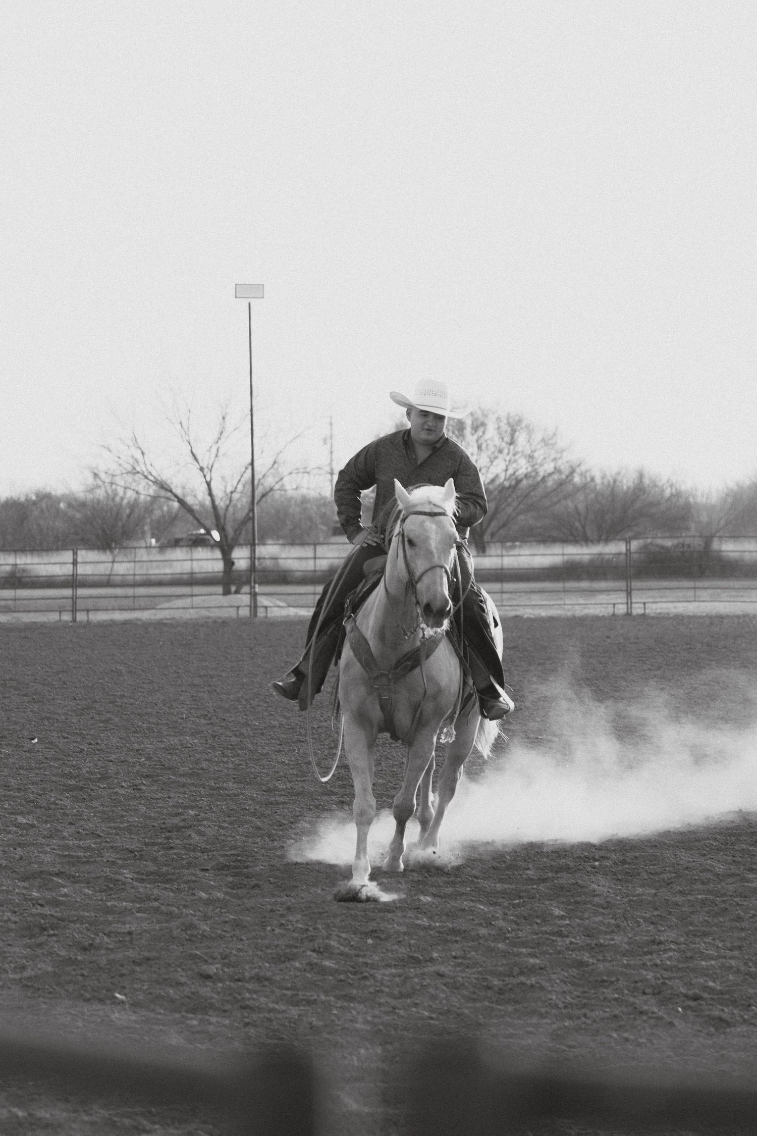 A monochrome image of a rider on horseback galloping across a dusty arena leaving a trail of dust behind them.