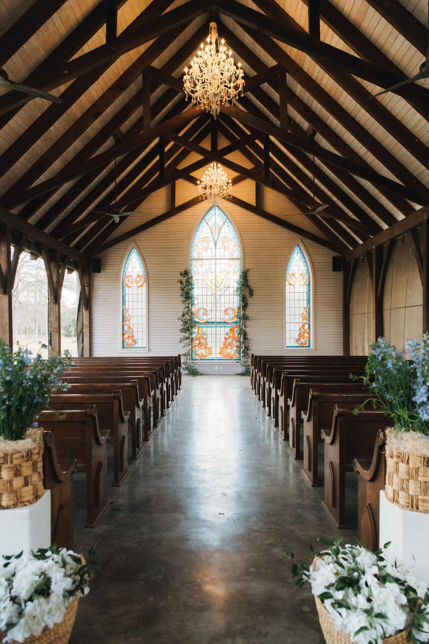 Interior view of a rustic chapel with wooden ceiling beams, stained glass windows and crystal chandeliers adorned with floral decor.