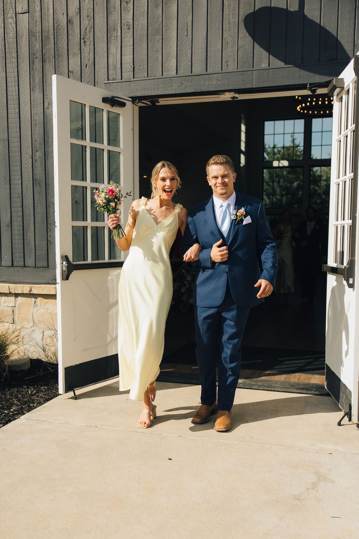 A couple joyfully exits their wedding venue through white french doors wearing a navy suit and cream gown.