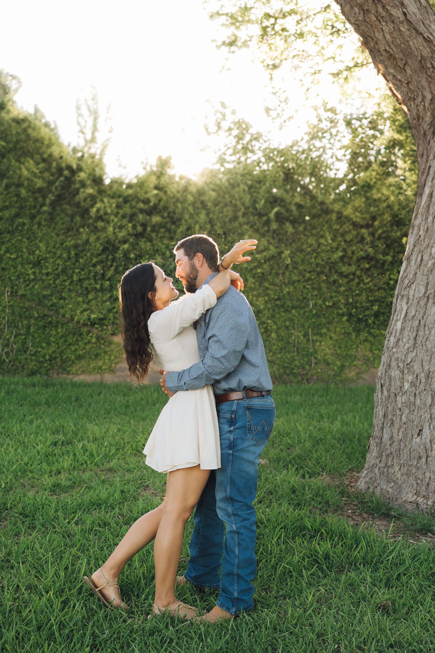 A romantic couple dances playfully together on green grass with trees and hedges in the background on a sunny day.
