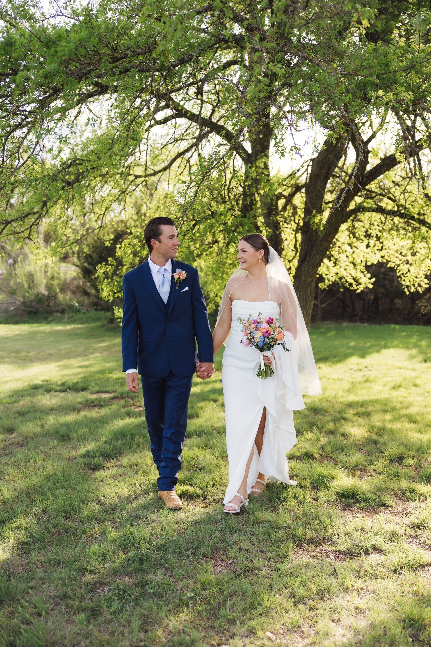 Couple walks together through a sun-dappled garden setting with willow tree branches overhead during outdoor wedding ceremony.