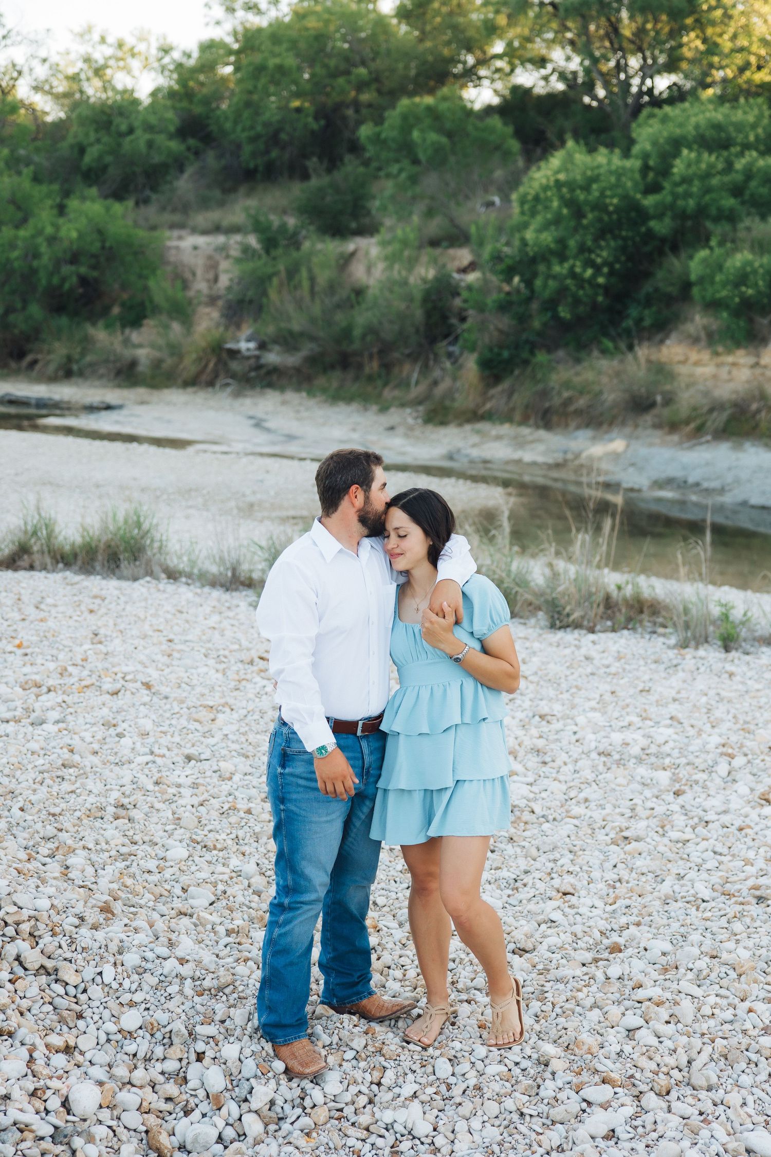 A romantic couple embraces on a rocky riverbank surrounded by lush greenery on a sunny afternoon.