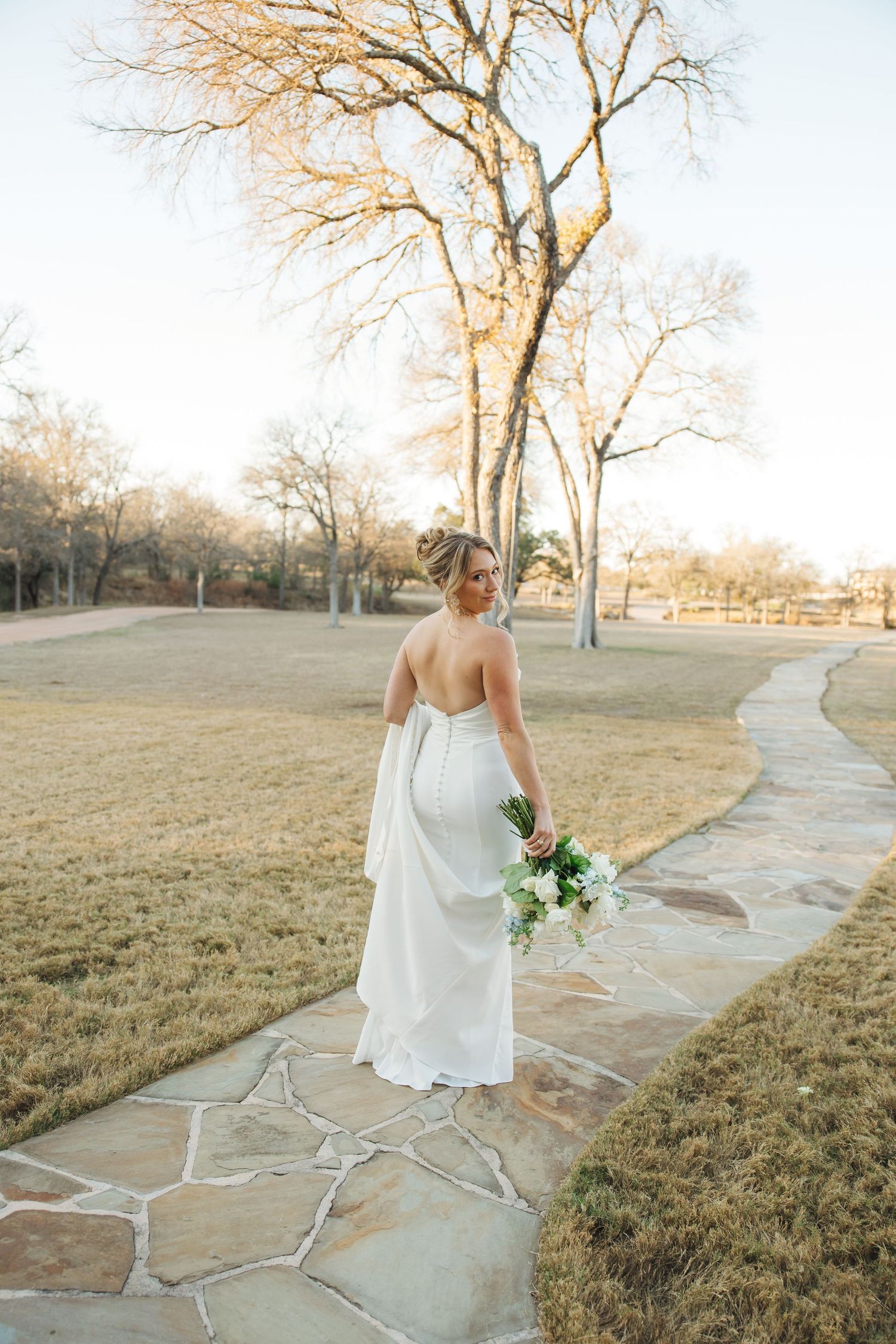 A bride in a white wedding dress walks along a winding stone path through a grassy field at golden hour.