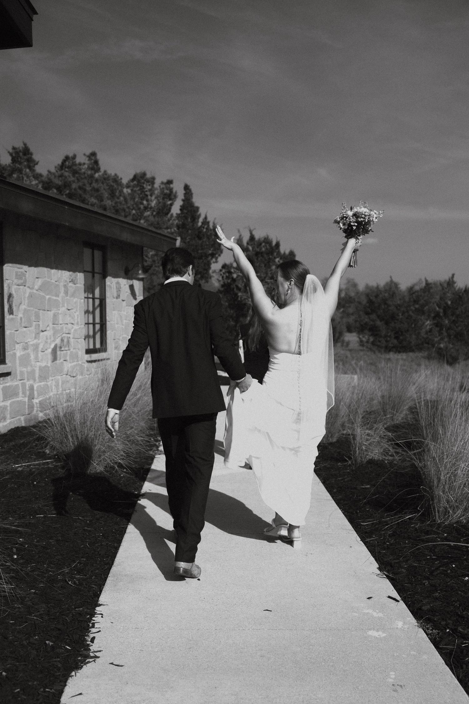 A black and white wedding photo series showing a couple walking together outdoors near a stone building.