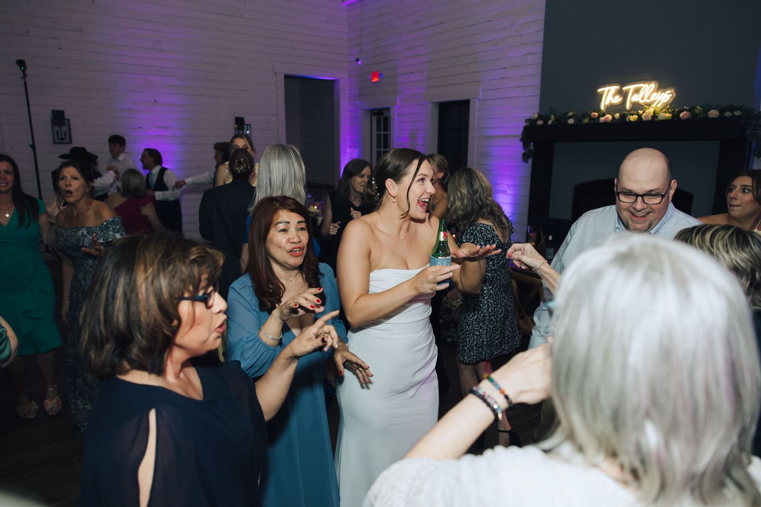 Wedding party guests dancing and celebrating on a crowded dance floor with purple uplighting in the background.