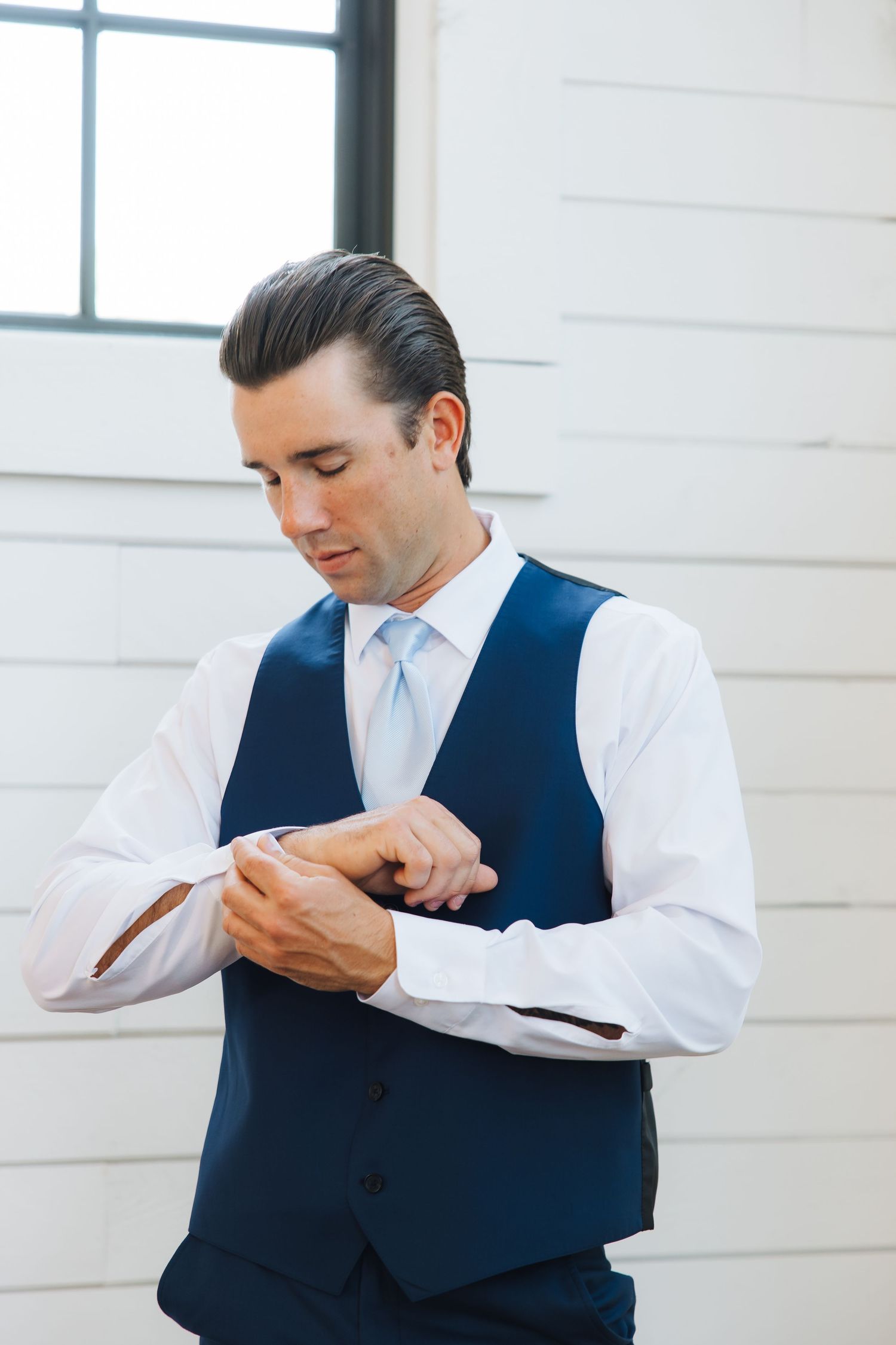 A person in a navy blue vest and white dress shirt checks their watch while standing against a light wall.