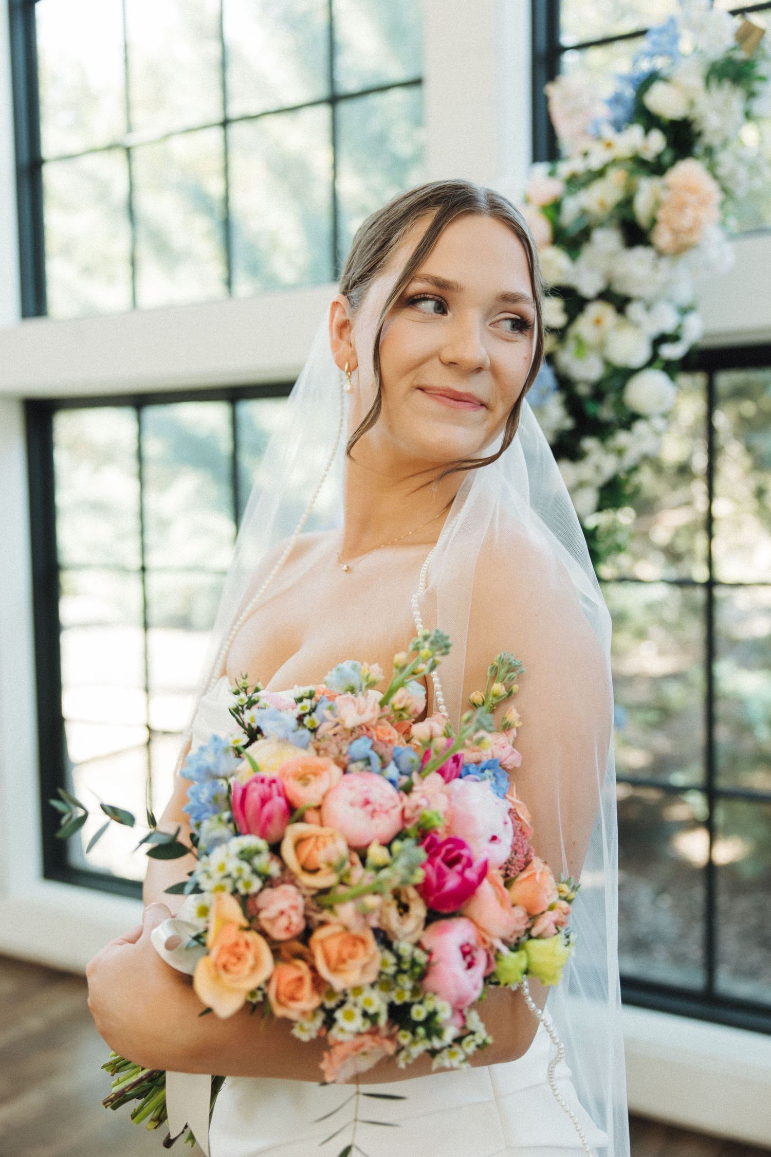 Colorful bridal bouquet features pink, orange, and coral roses with mixed florals against sunny window backdrop.