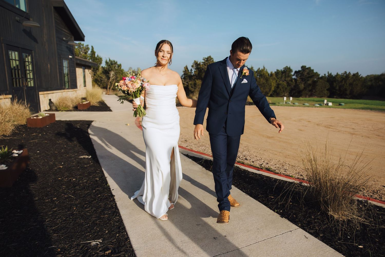 A couple holds hands while walking along a concrete path outside a rustic barn venue on their wedding day.