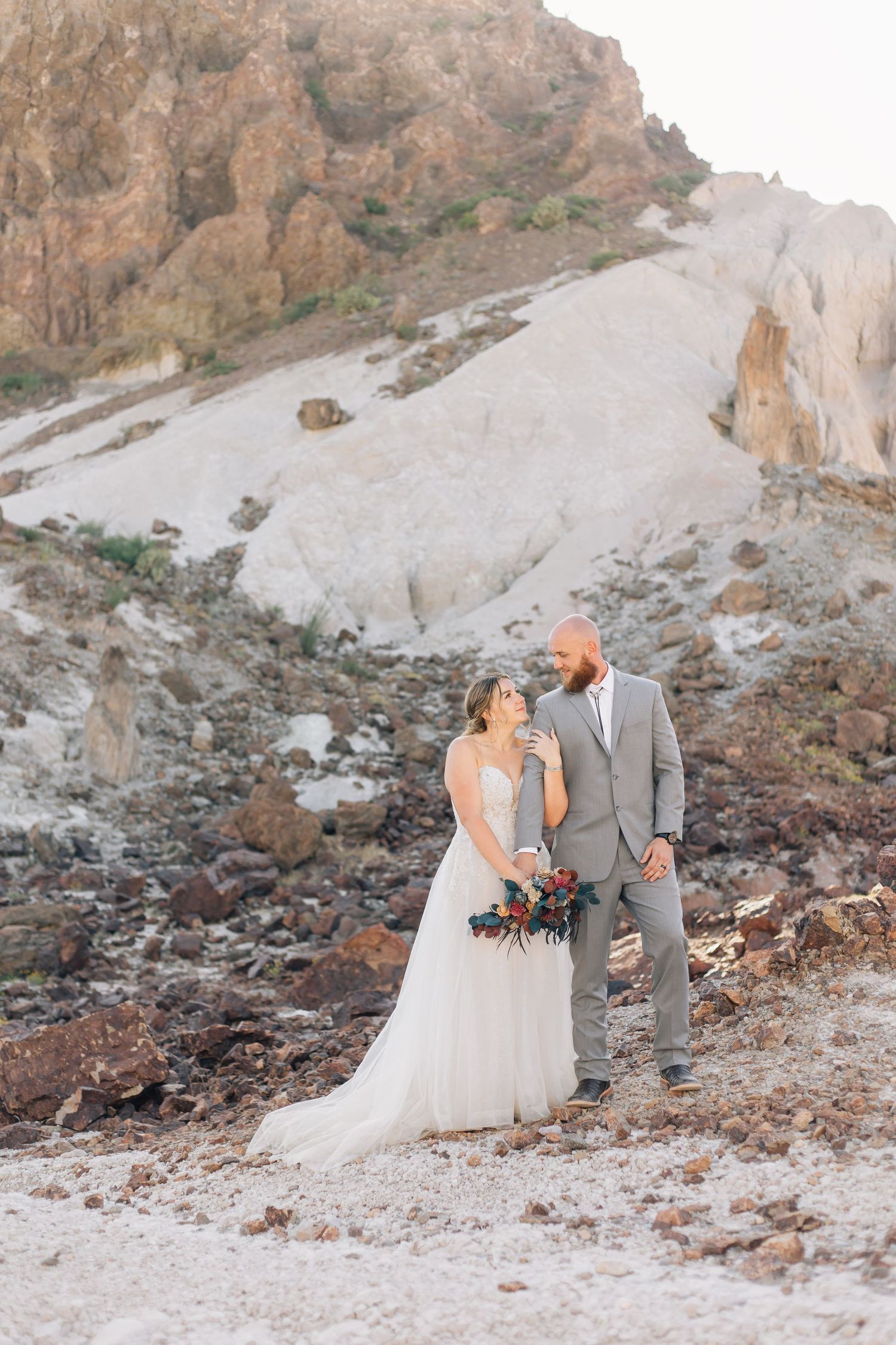 A couple in wedding attire standing on rocky mountainous terrain with a white cliff face in the background.