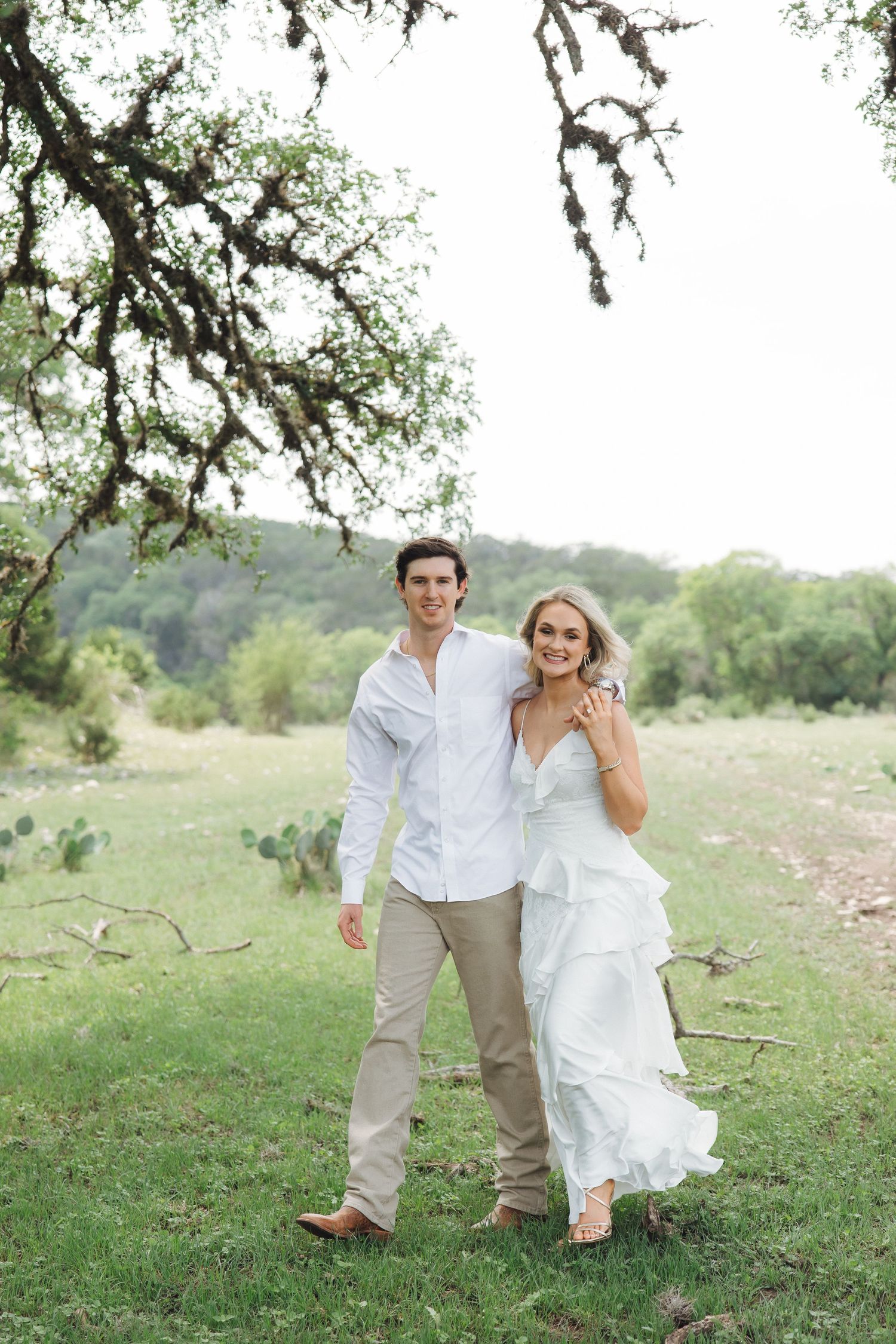 A couple runs playfully through a grassy field under a tree branch on a warm sunny day.