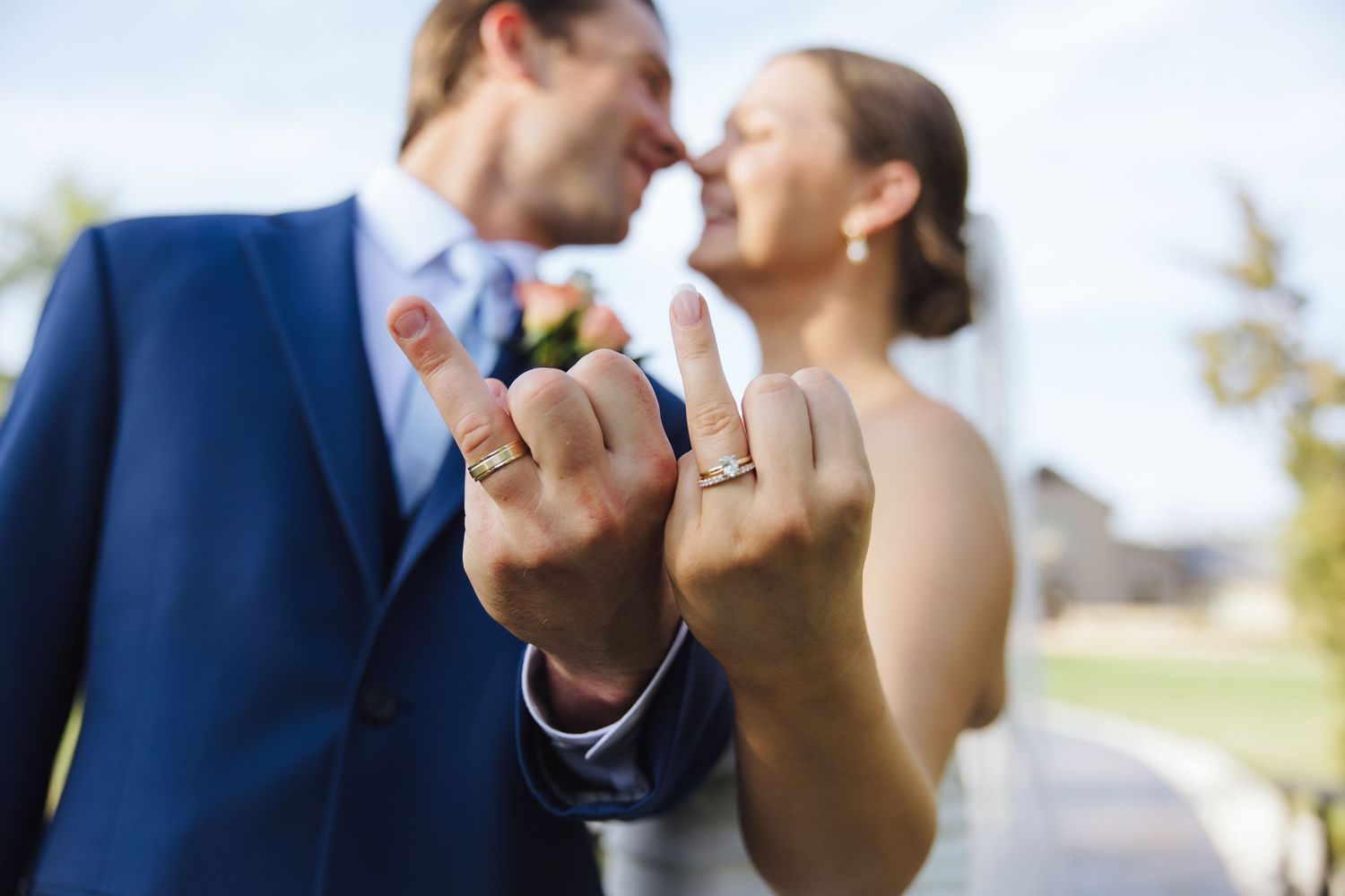 Closeup of hands displaying wedding rings while couple shares an intimate moment outdoors.
