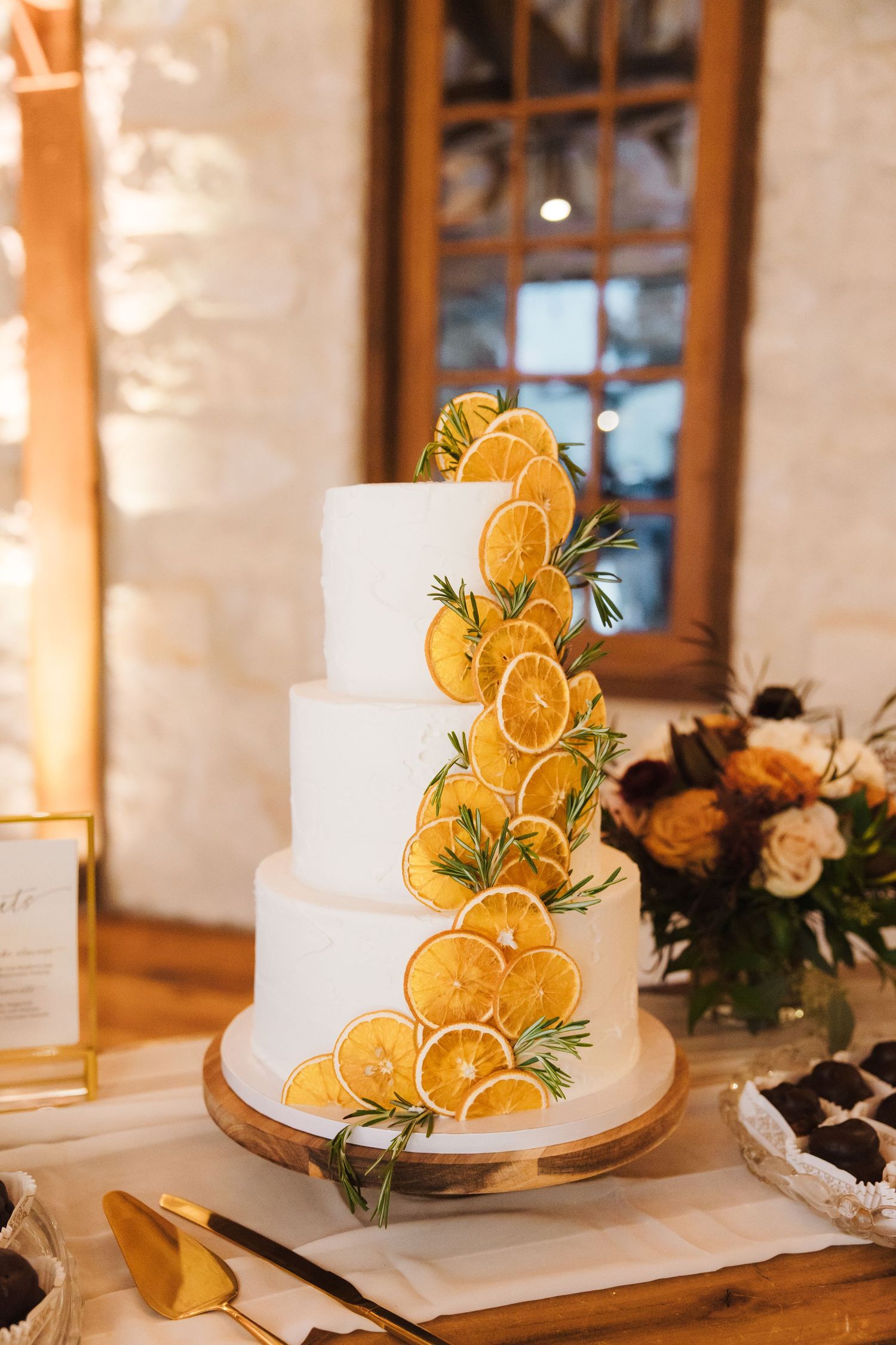 A wedding cake decorated with dried orange slices and fresh rosemary sits on a rustic wooden cake stand.