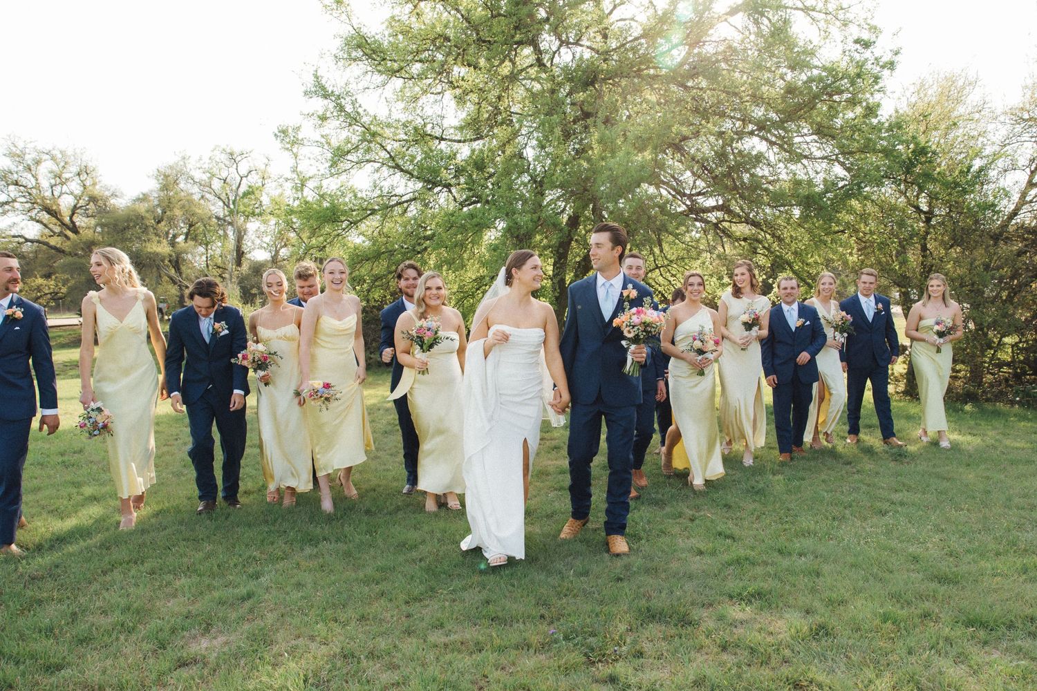 Wedding party lined up outside on grassy hill wearing navy suits and blush bridesmaid dresses for formal group photo.