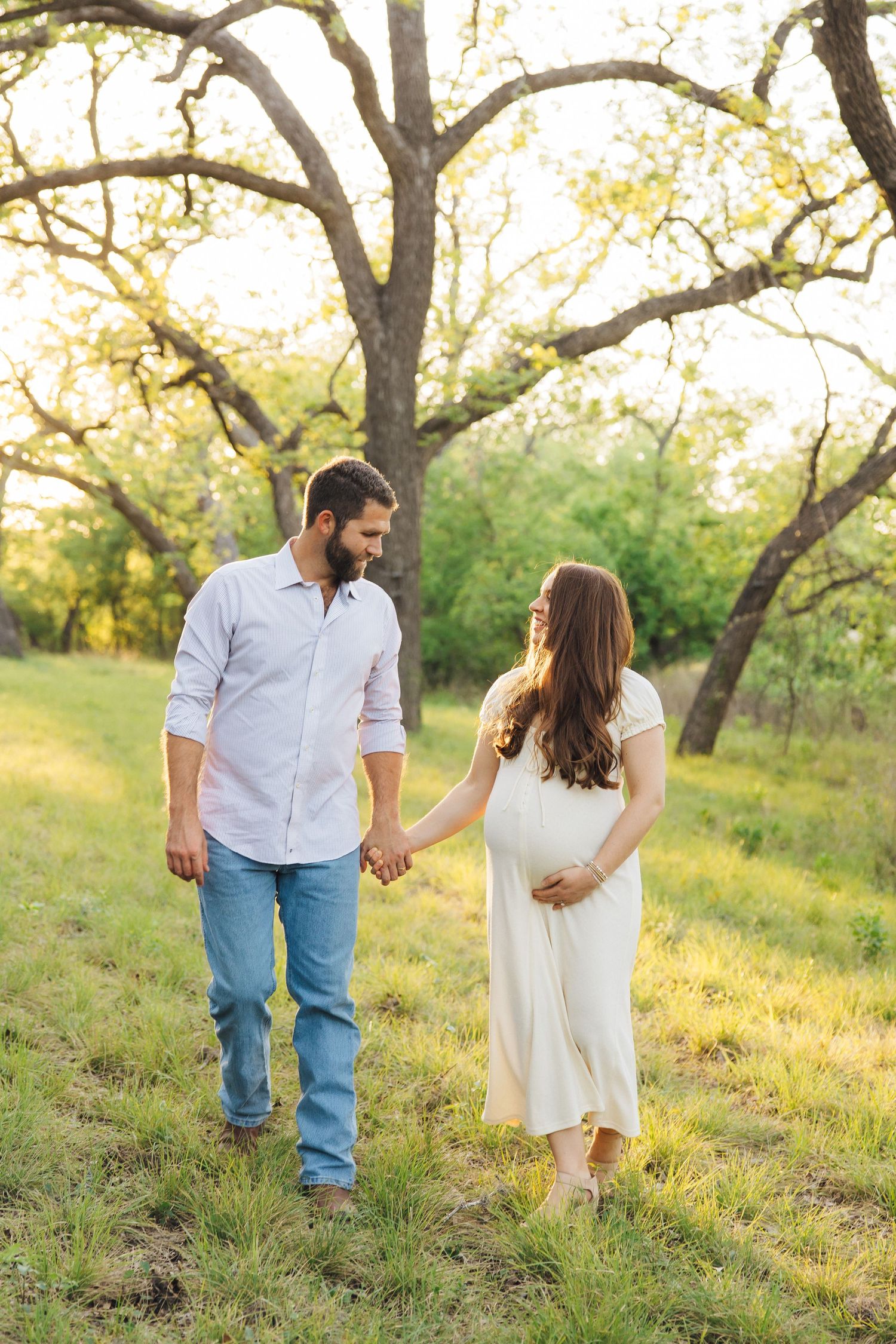 A couple holding hands walks together on a grassy hillside beneath large trees during golden hour.