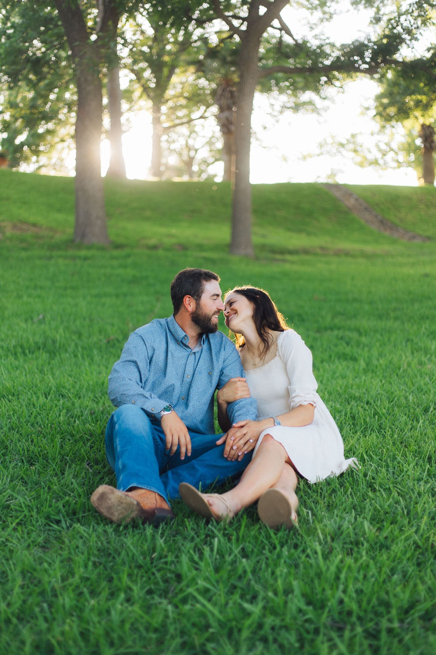 A couple sits together on green grass during sunset, sharing a tender moment beneath trees in golden light.