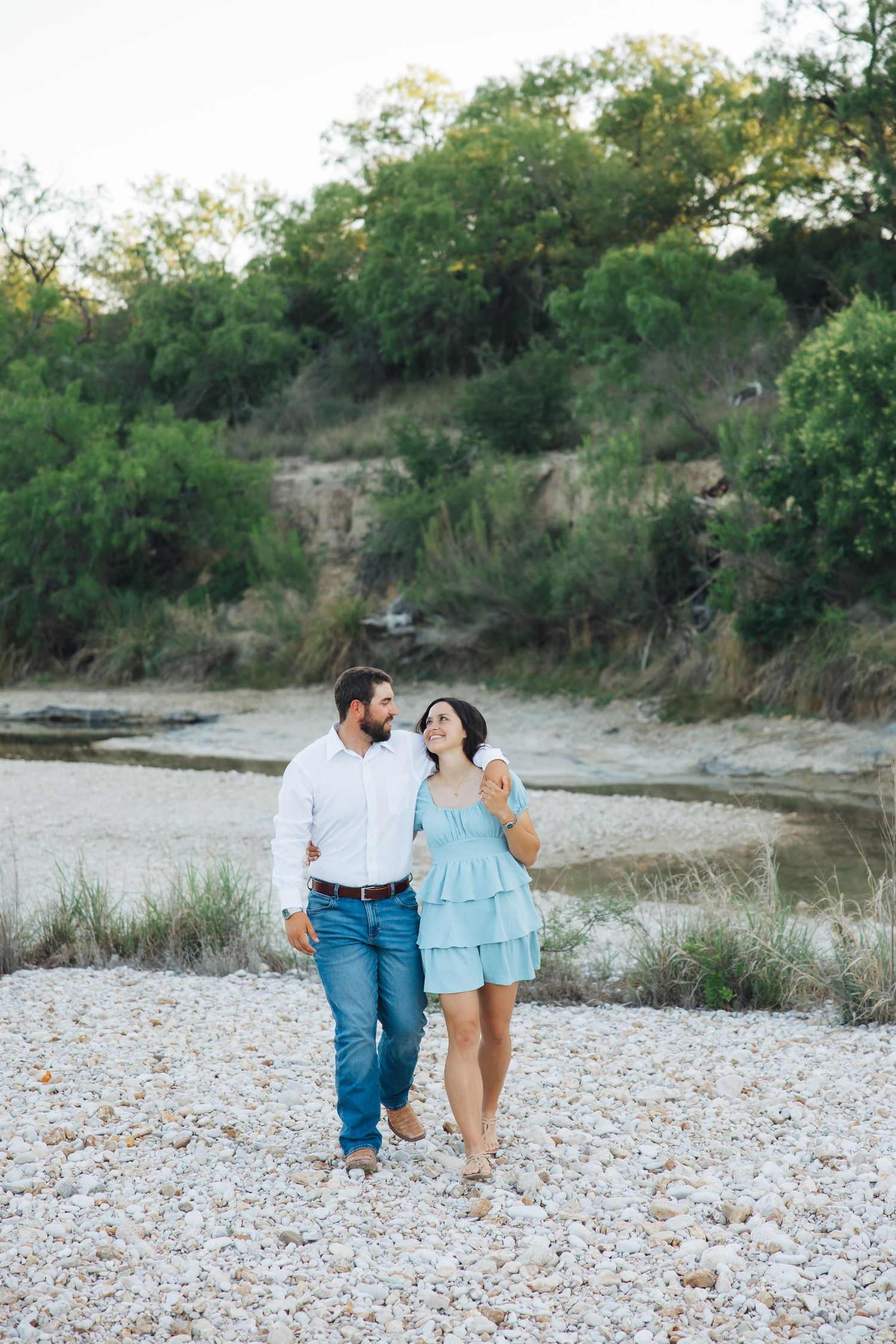 A couple walks together along a rocky riverbank with cliffs and greenery in the background.