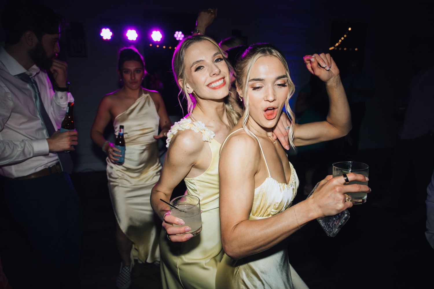Group of party-goers dance together on a dance floor with colorful purple lighting in a nightclub setting.