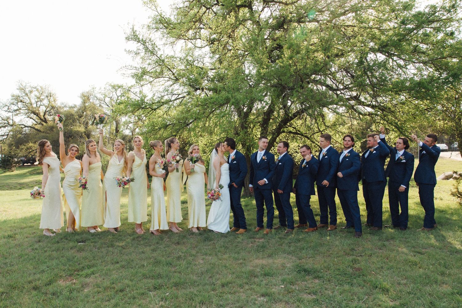 Wedding party in yellow bridesmaid dresses and navy suits poses together on a grassy lawn under large trees.