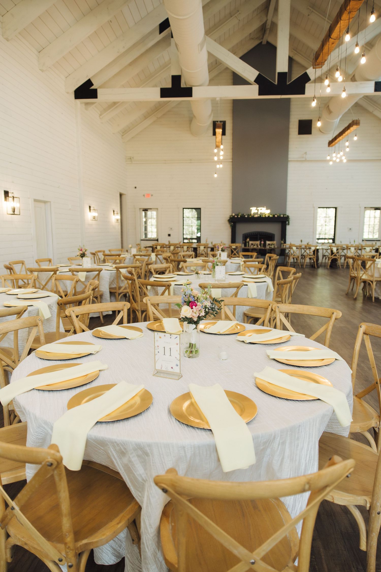 White and light gray wedding reception venue with vaulted ceiling, round dining tables and wooden cross-back chairs.