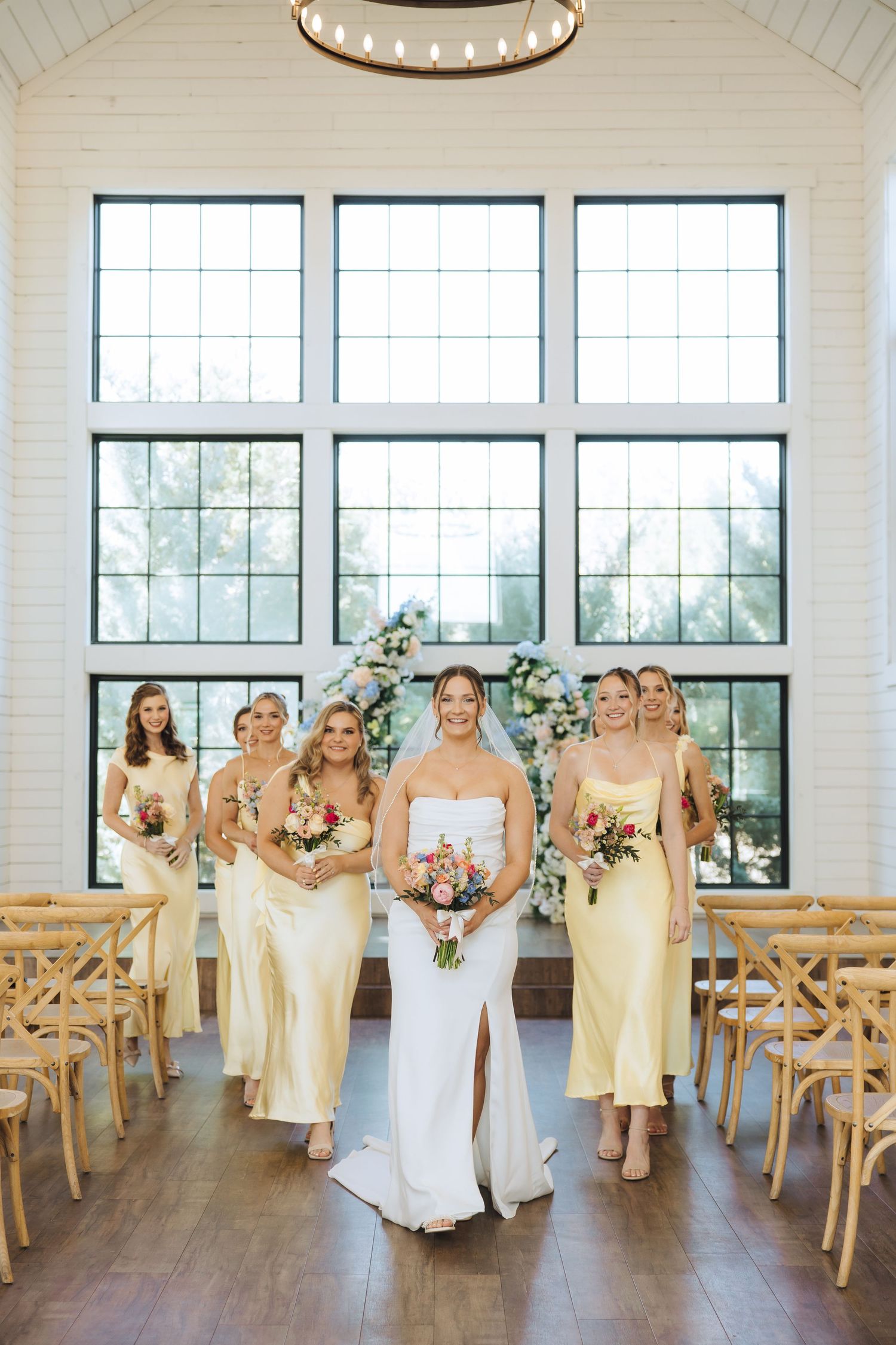 Wedding party in soft yellow dresses and white gown gathered in a bright modern venue with wooden chairs.