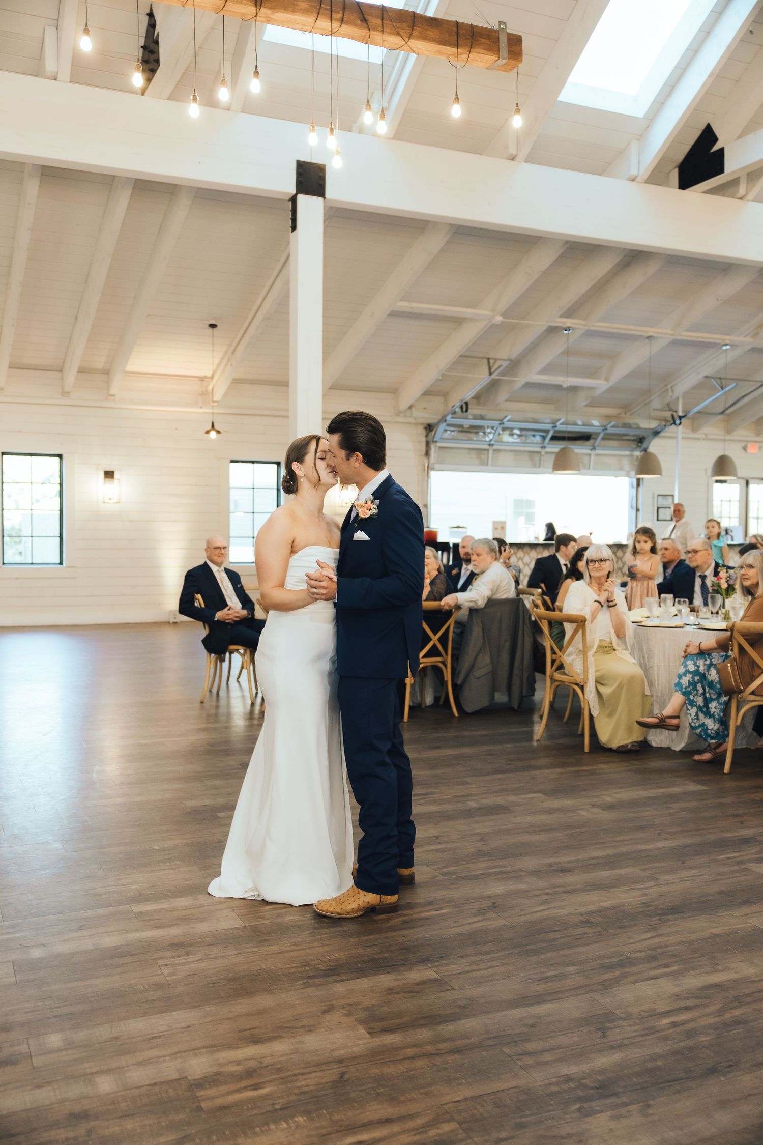 A wedding couple shares their first dance in an elegant white venue with wooden floors and hanging pendant lights.