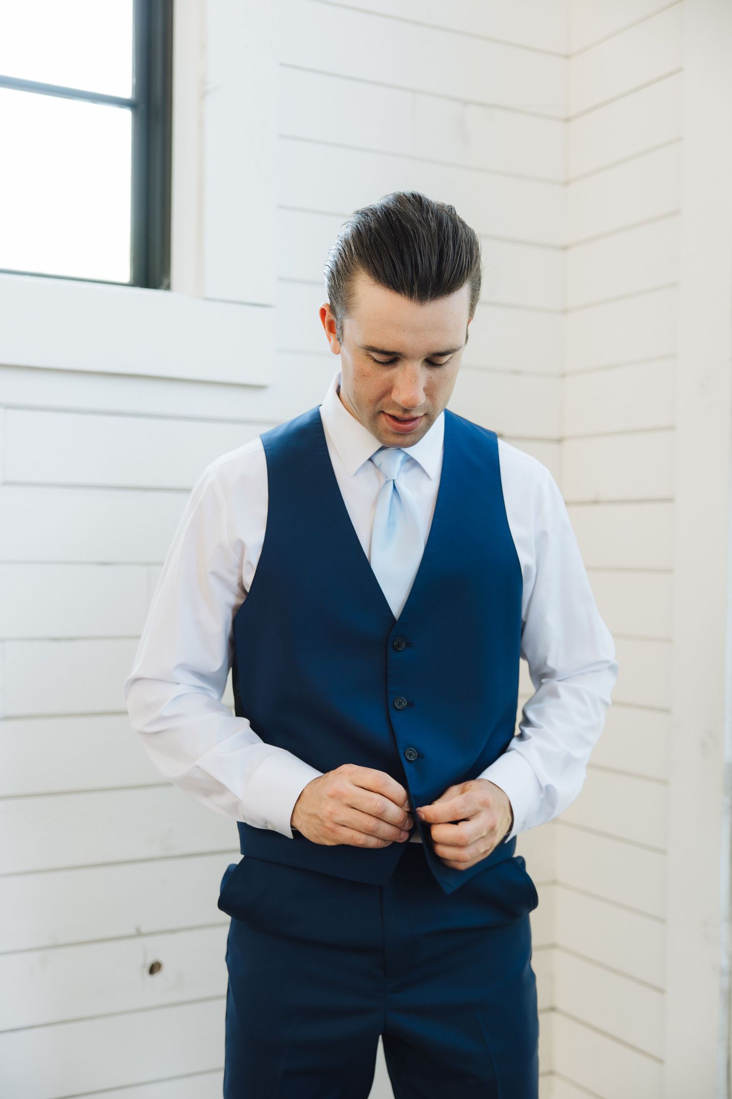 A person in a formal navy blue vest and white dress shirt buttons up their waistcoat while standing in a corner.