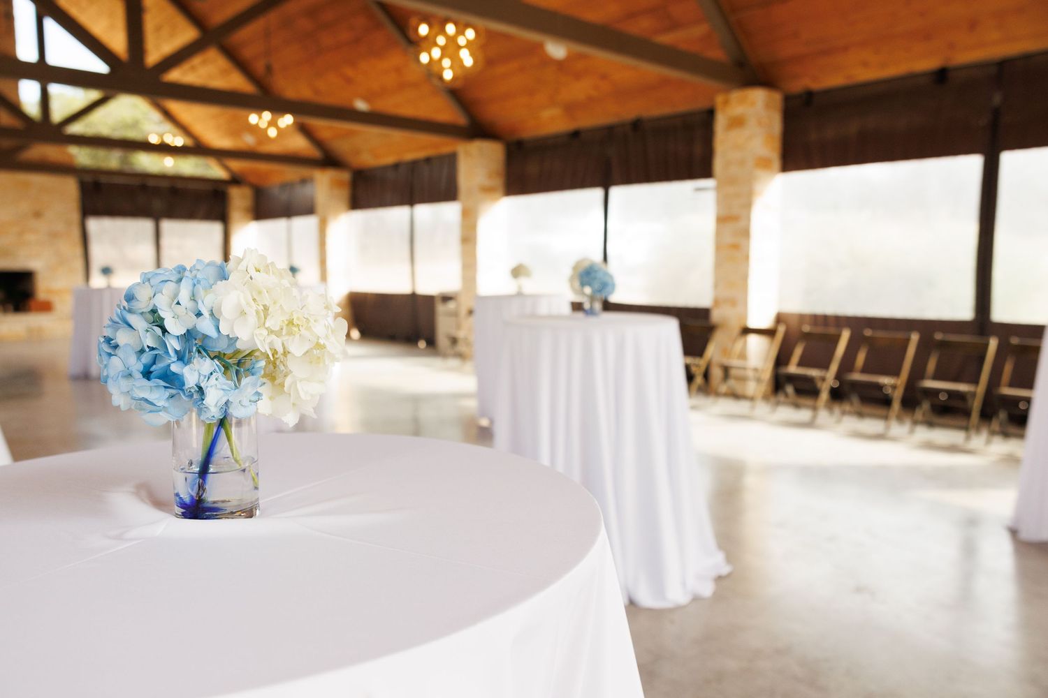 Wedding reception venue with white tables, light blue floral centerpieces and wooden beam ceilings in rustic space.
