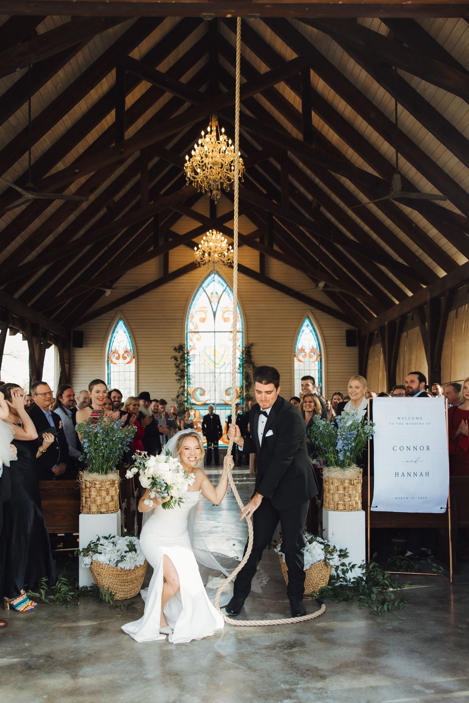A celebration in a rustic chapel with exposed wooden beams, stained glass windows, and an elegant crystal chandelier hanging above.