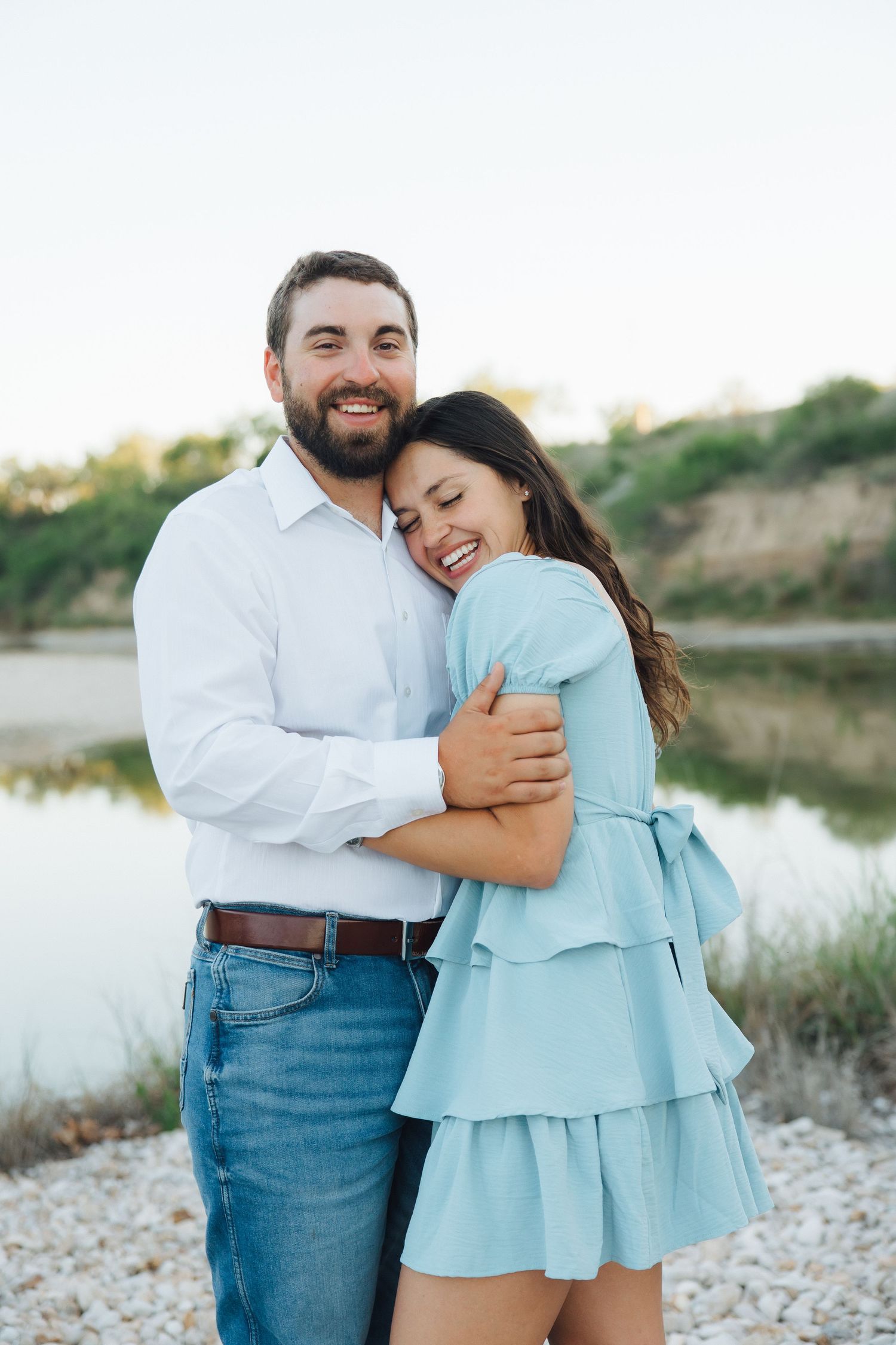 A romantic couple embraces by a serene lake at sunset, with the woman wearing a light blue ruffled dress.