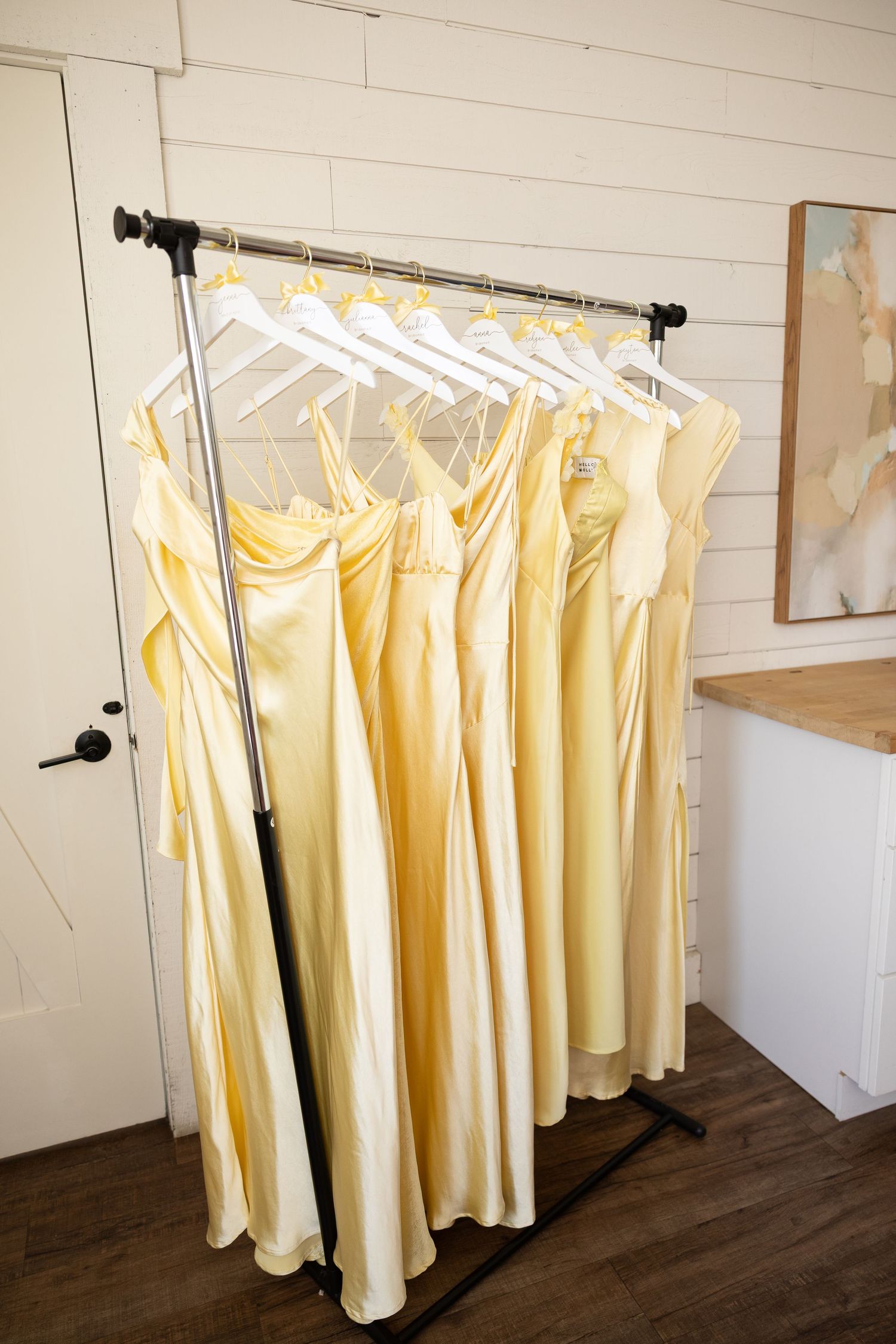 Several pale yellow bridesmaid dresses hanging in a row on white hangers against a white wall.