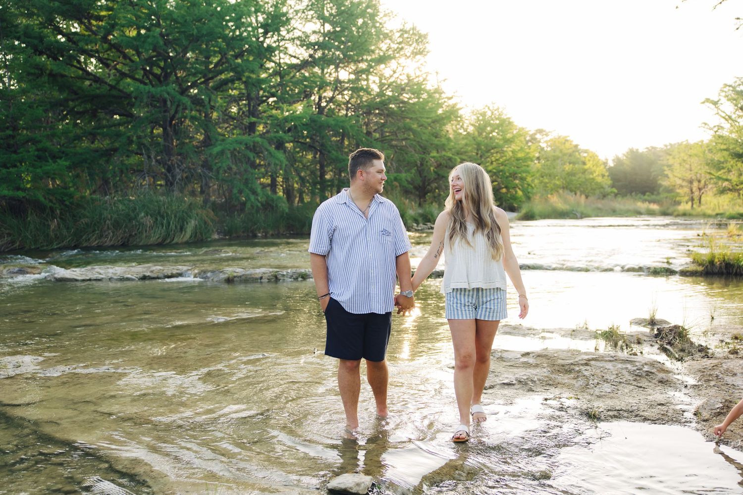 A couple wading in shallow water hold hands while walking along a scenic riverbank during sunset.