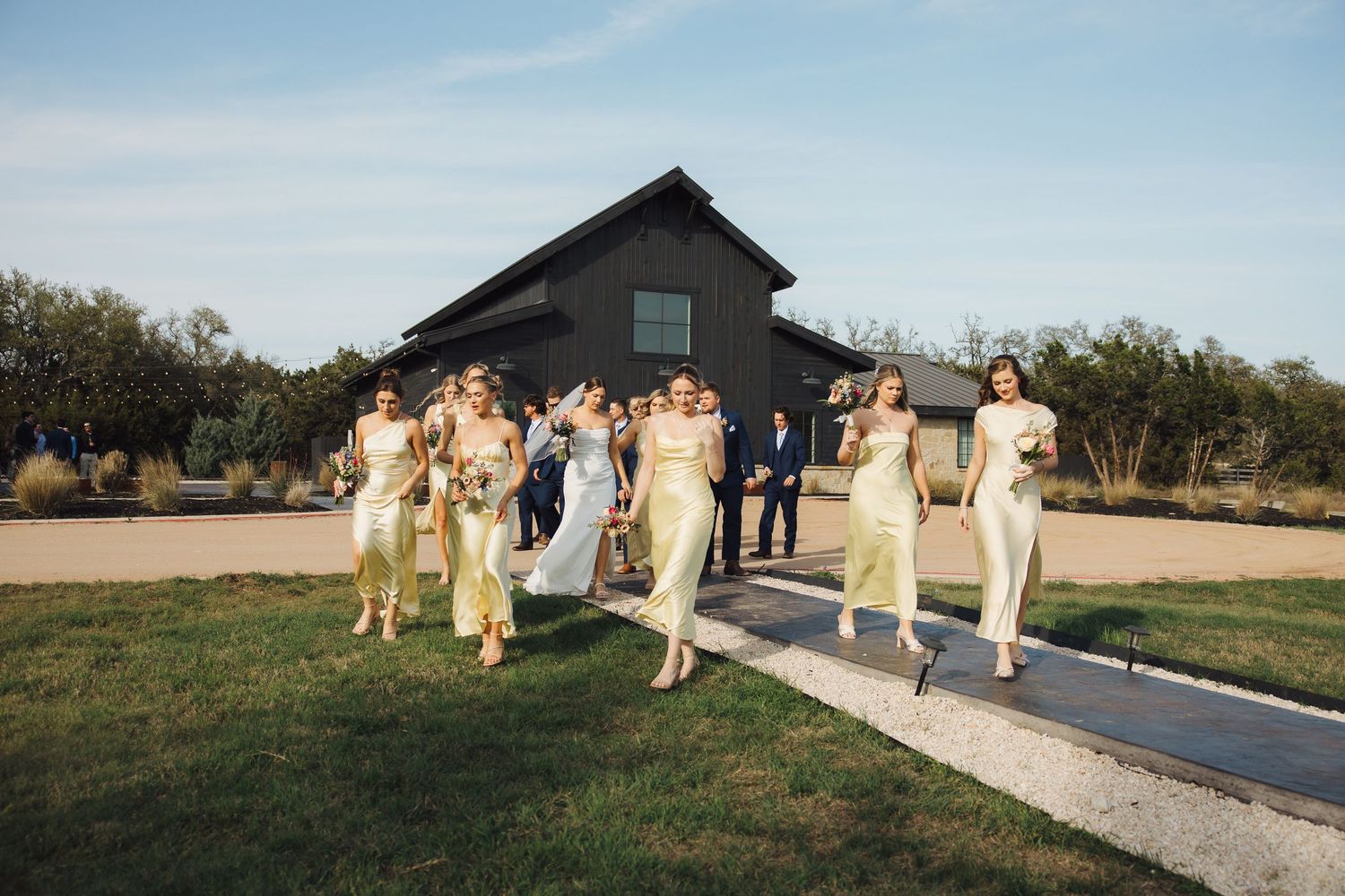 A bridal party dressed in cream yellow dresses walks down an outdoor path at a countryside venue at dusk.