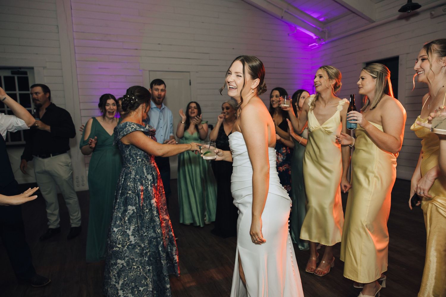Wedding guests in yellow and blue dresses dance and celebrate together in a reception venue with purple lighting.