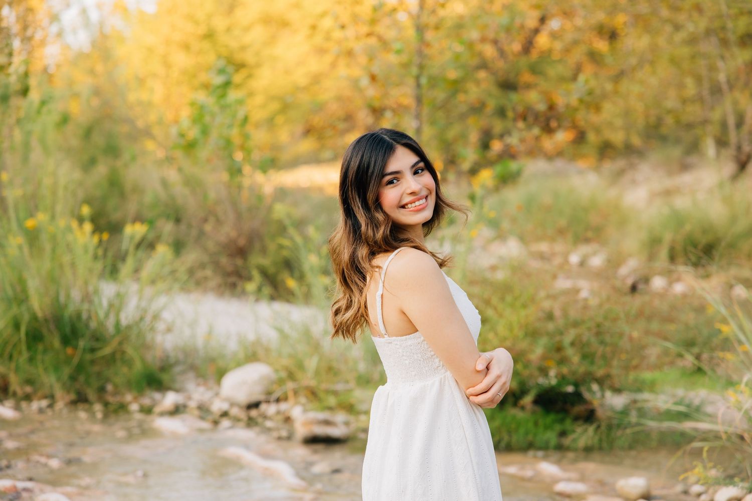 A woman in a white dress poses by a stream during autumn with golden foliage in the background.