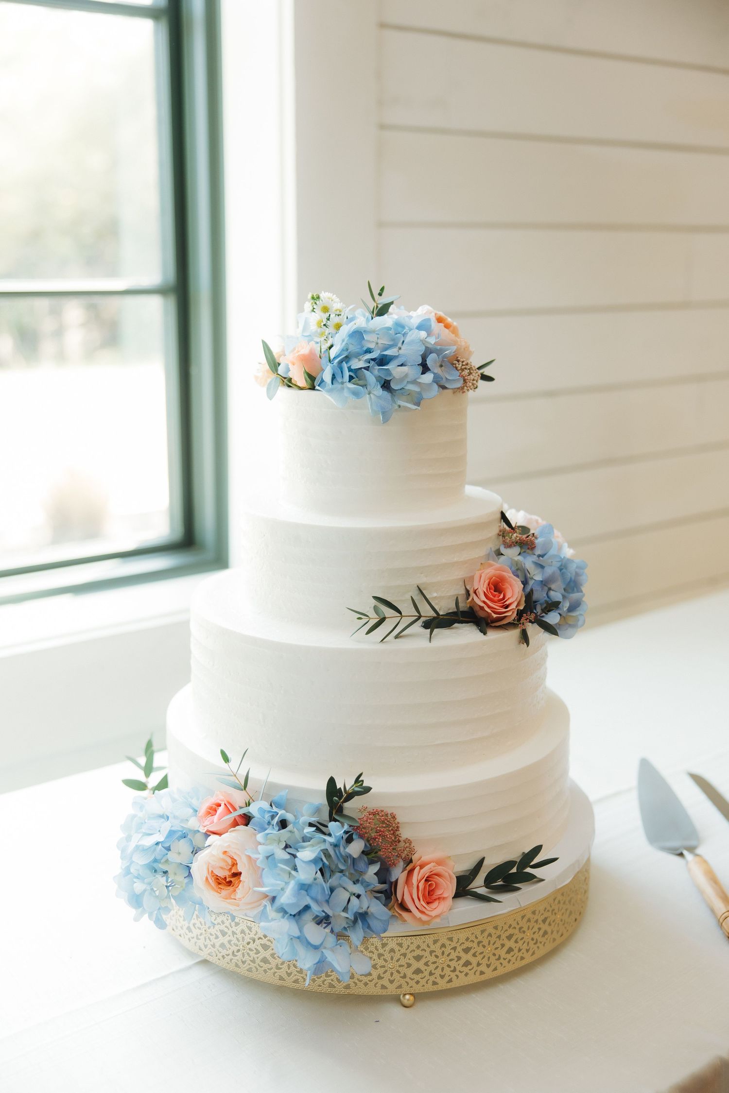 Elegant white wedding cake decorated with light blue hydrangeas and peach roses against a white shiplap wall.