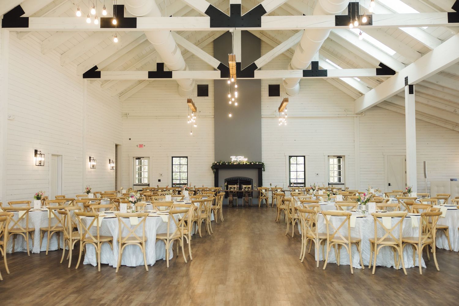 Elegant white barn wedding venue interior with exposed beams, round tables, and rustic wooden chairs set for reception.