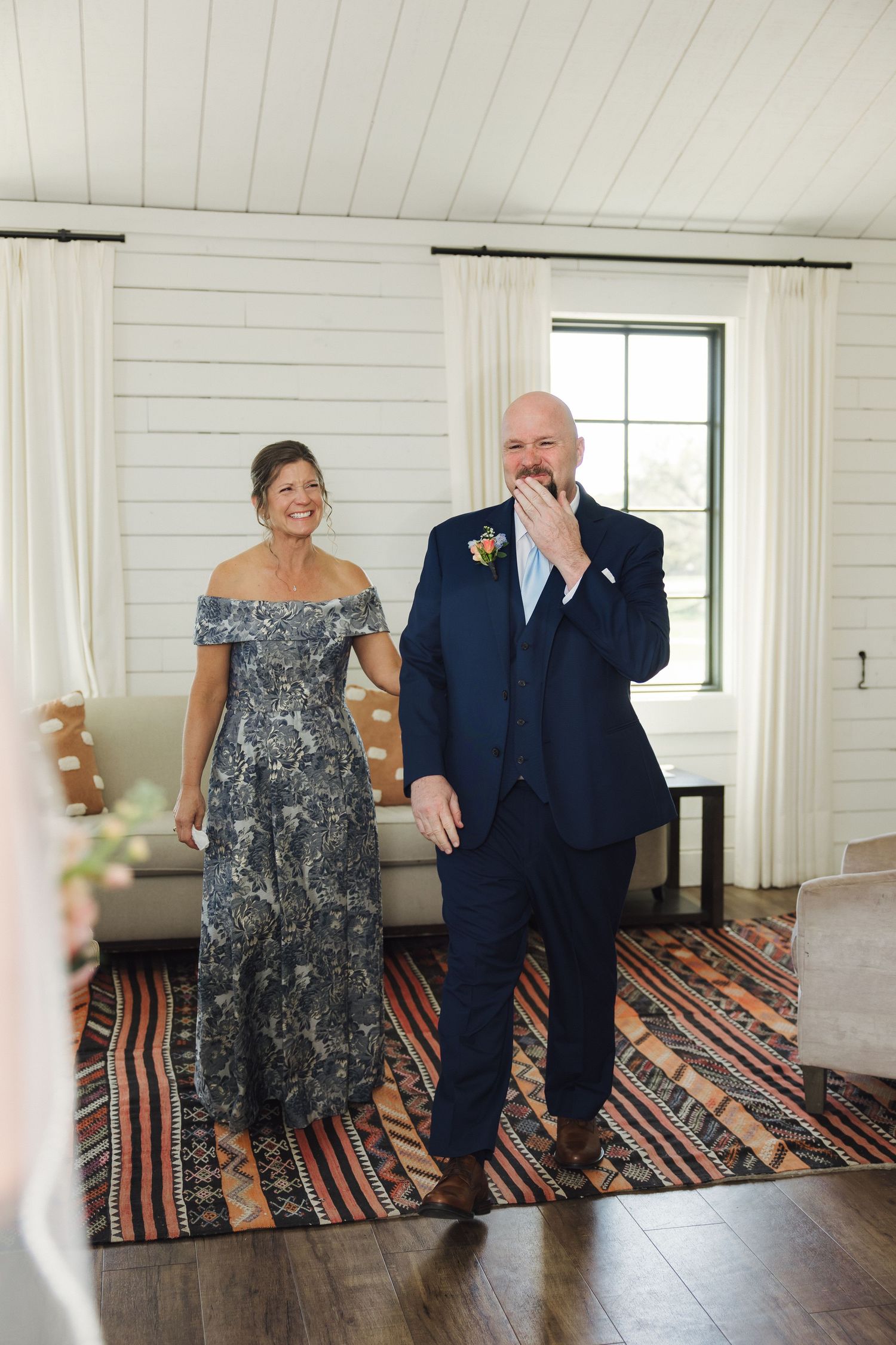 A couple shares an emotional first look moment in a white-walled room with a striped rug.