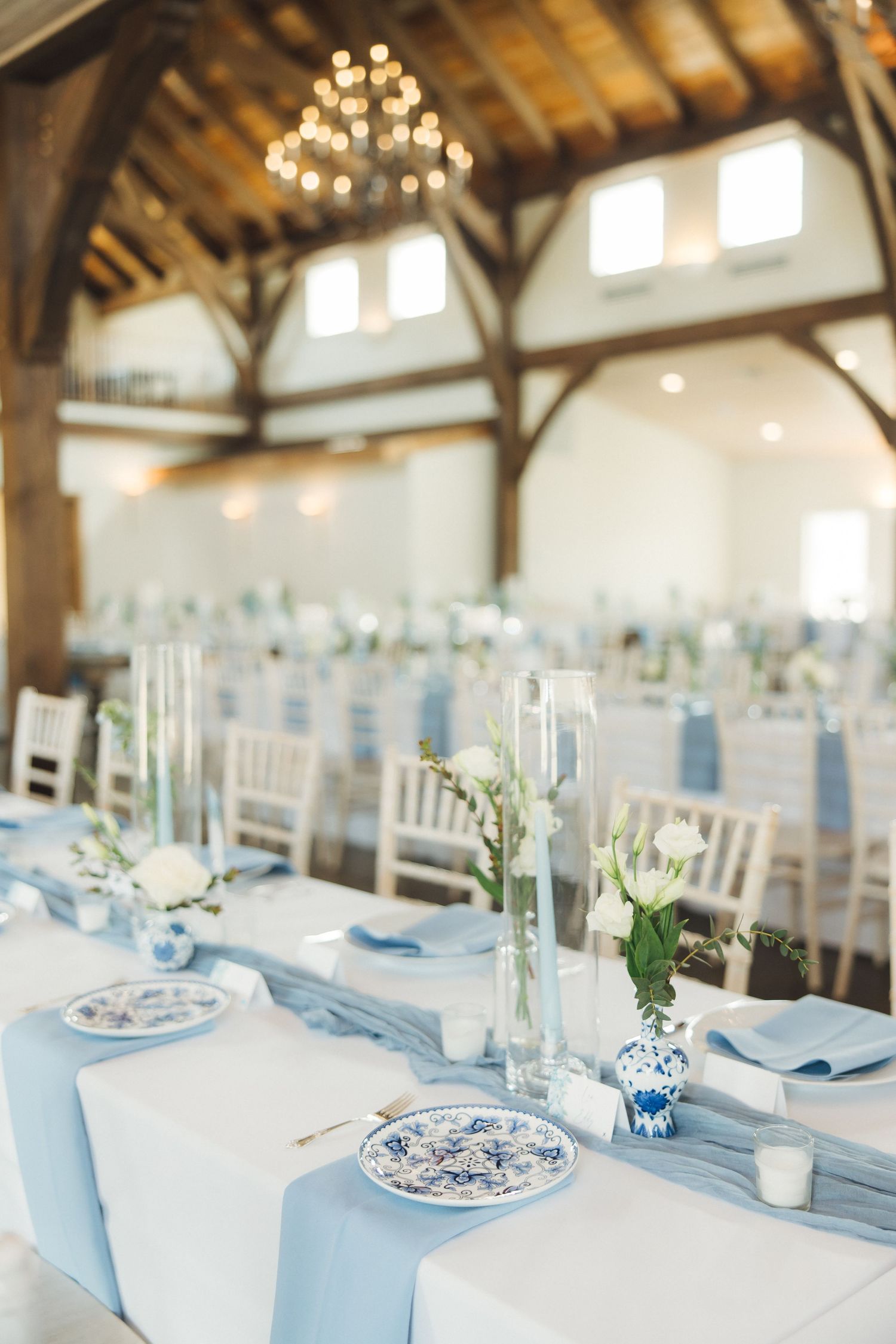 Elegant wedding reception venue with white and light blue table settings, china plates, and a rustic wooden beam ceiling.