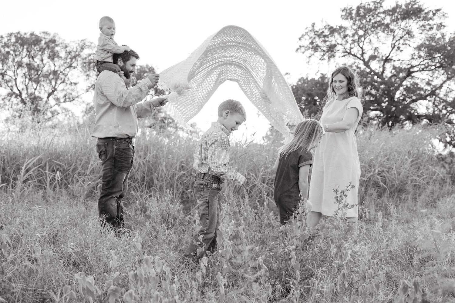 A black and white photo of a family playing with a flowing fabric in a grassy meadow on a sunny day.