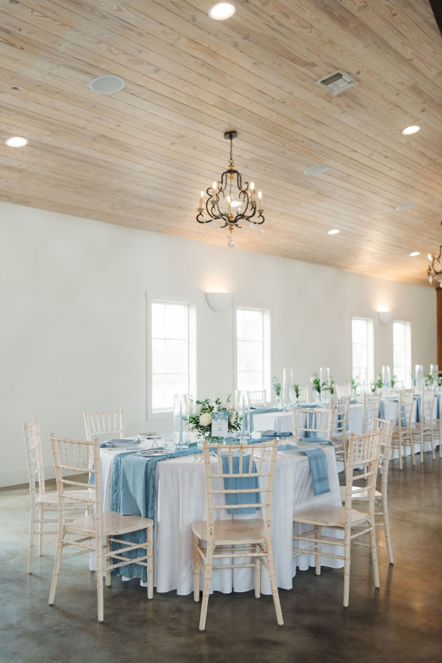 Elegant wedding reception venue with white chiavari chairs, light blue linens, and a rustic wood plank ceiling with chandelier.