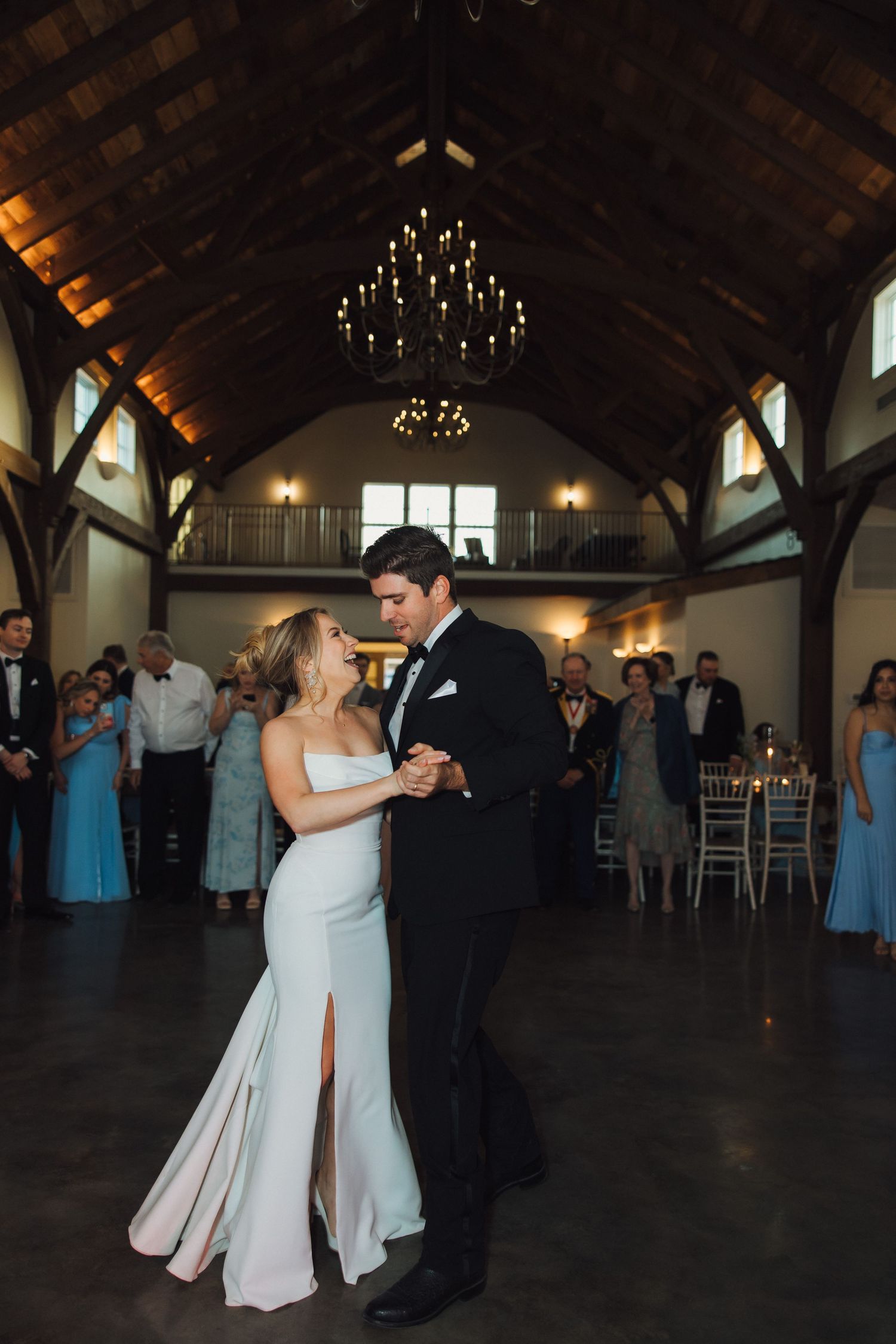 A couple shares their first dance in an elegant rustic venue with high wooden beams and chandeliers.